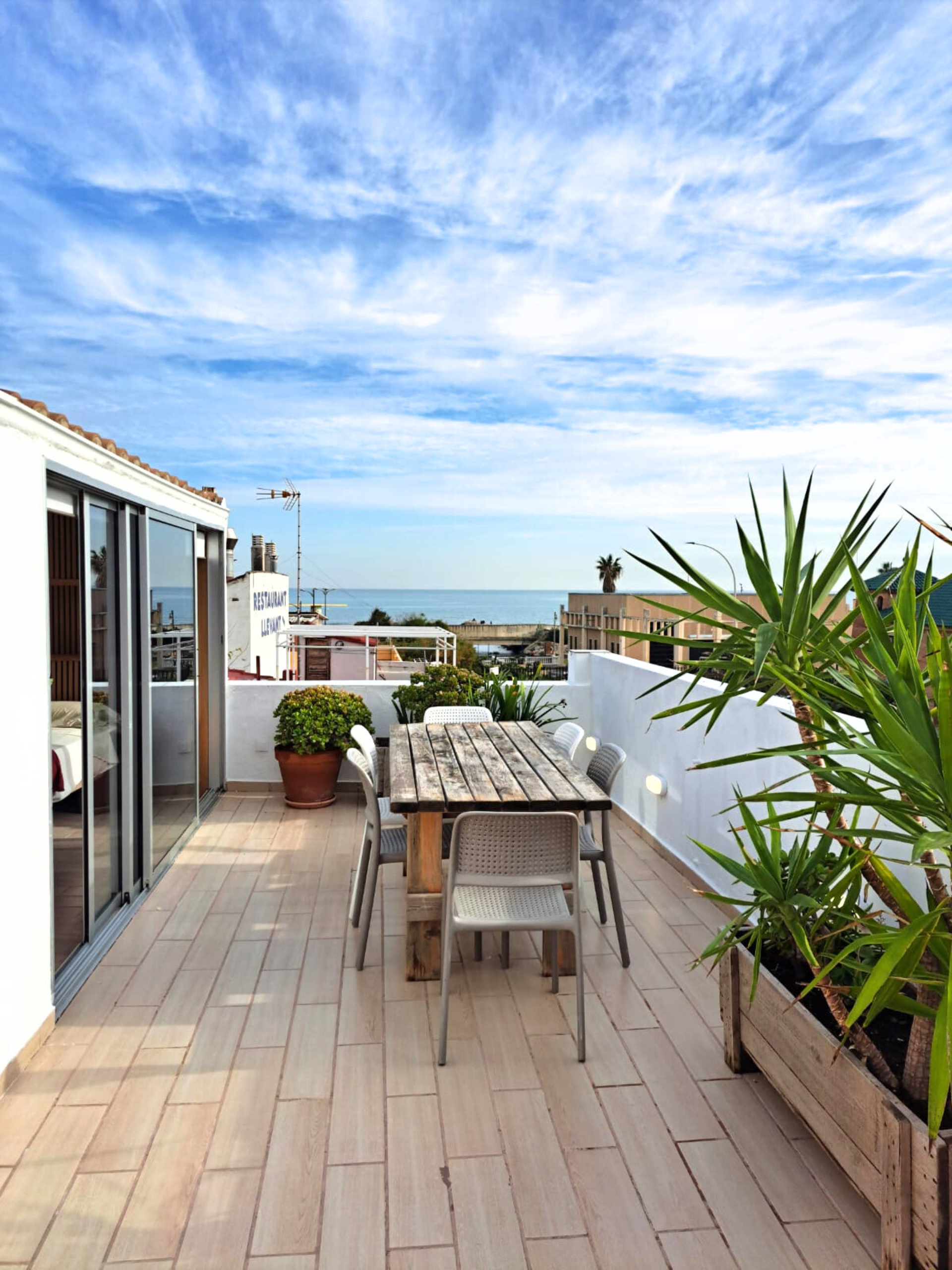 A wooden dining table and chairs on a tiled outdoor terrace overlooking the sea under a bright, blue, cloudy sky.