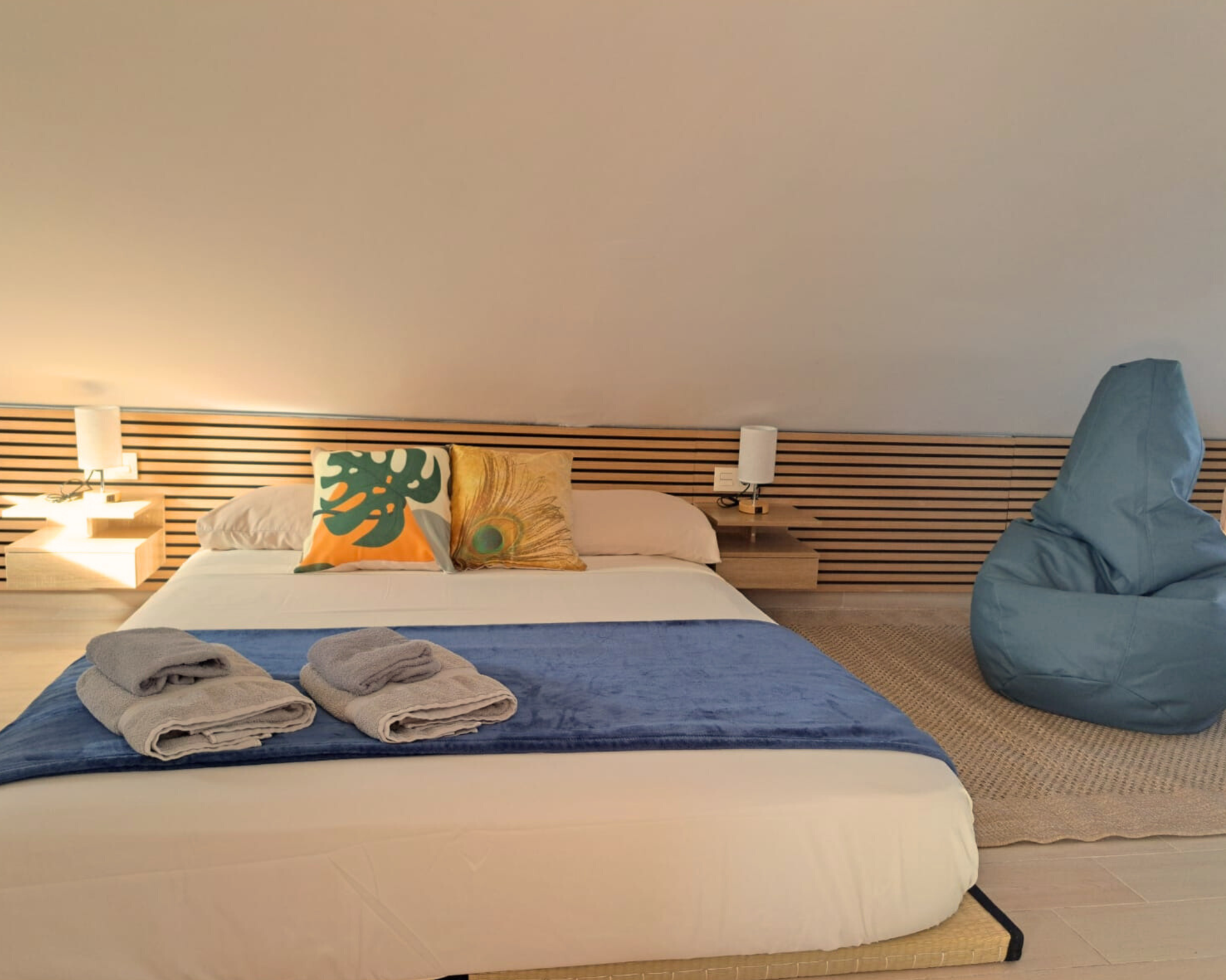 A modern bedroom with a low bed featuring blue bedding and decorative pillows, plus a blue beanbag chair in the corner.