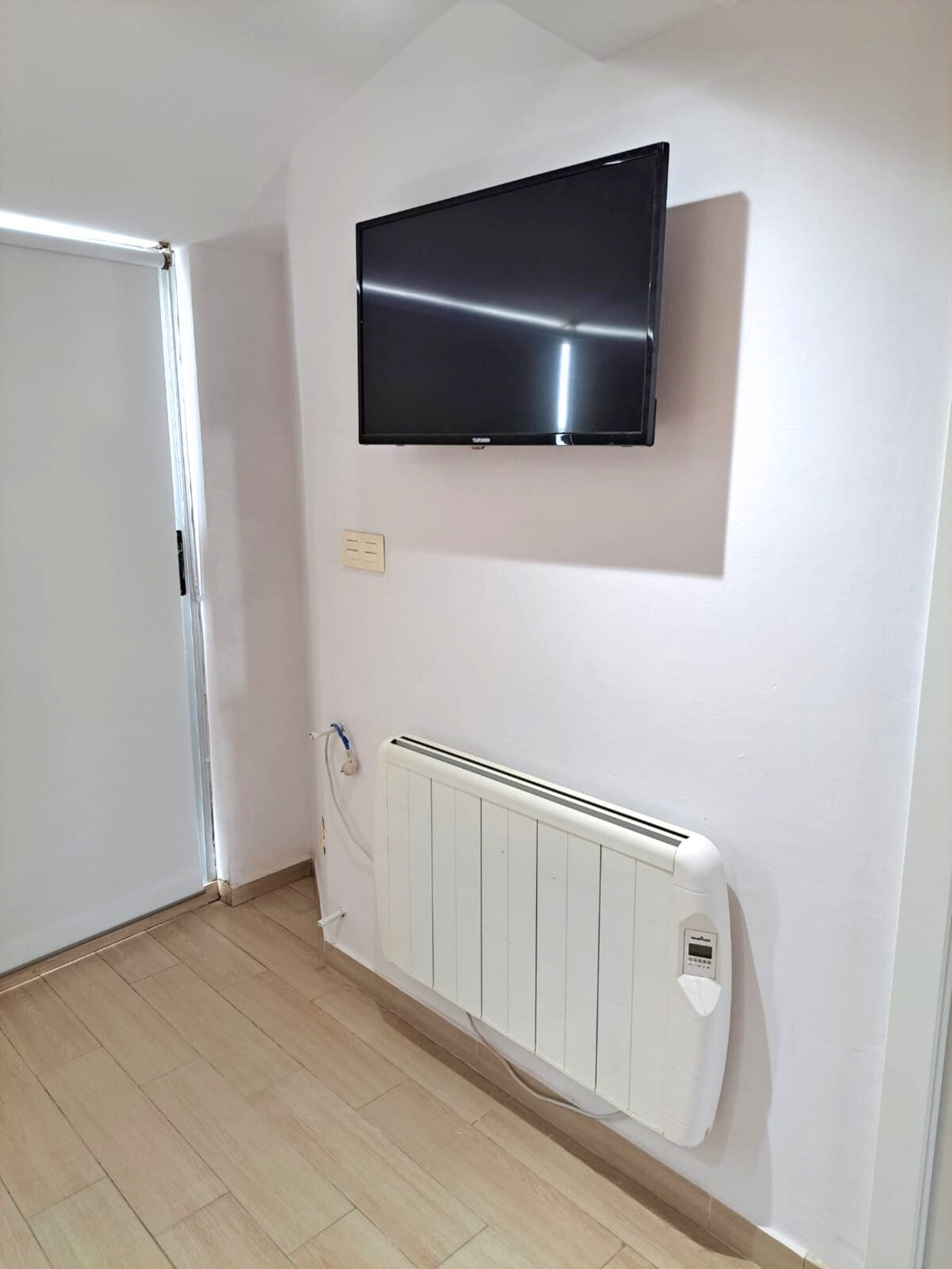 A flat-screen TV mounted on a light-colored wall above a white electric radiator in a room with light wood-look flooring.