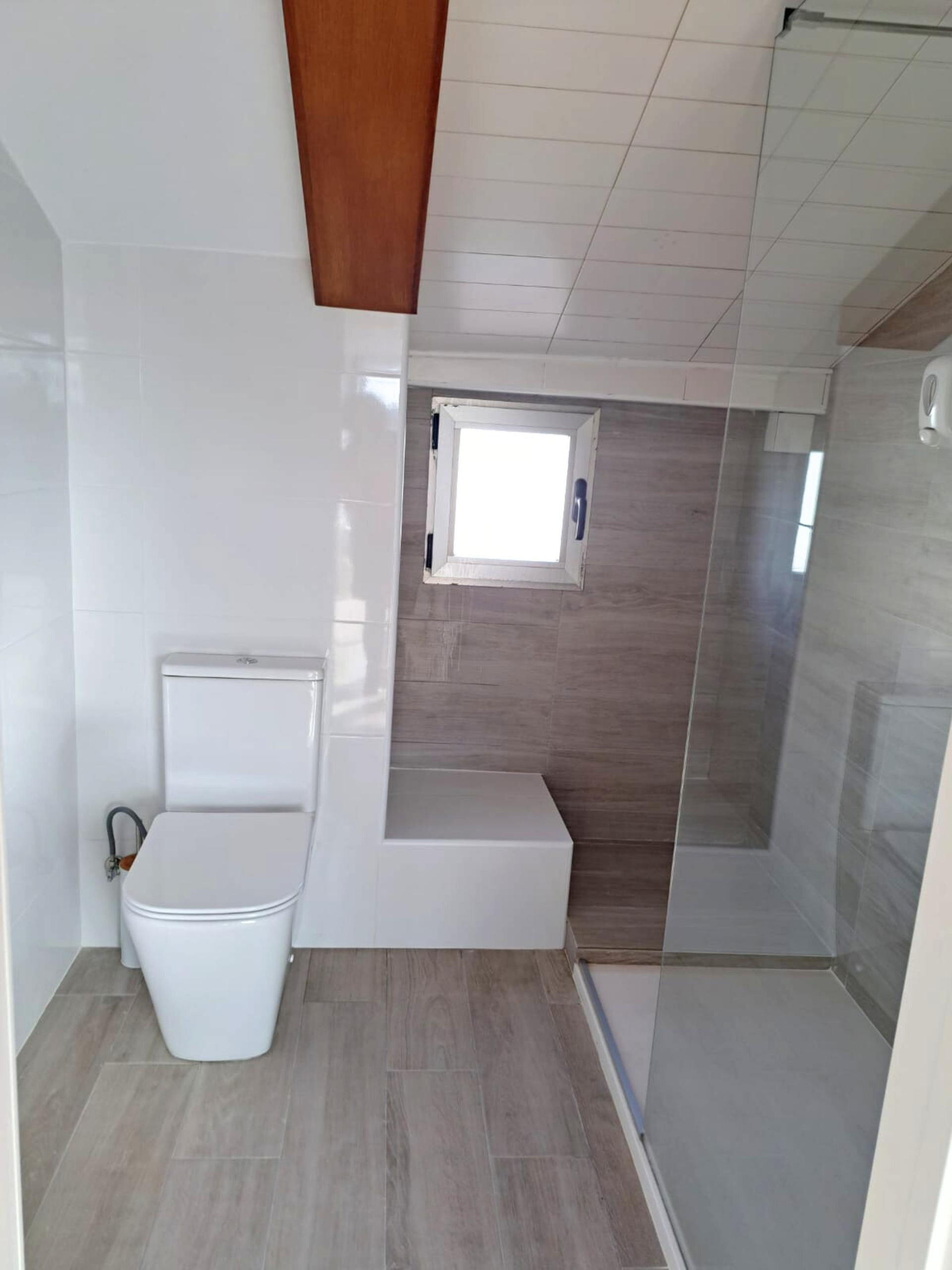 Modern bathroom with a white toilet, light wood-look floor tiles, a glass-enclosed shower, and a small window.
