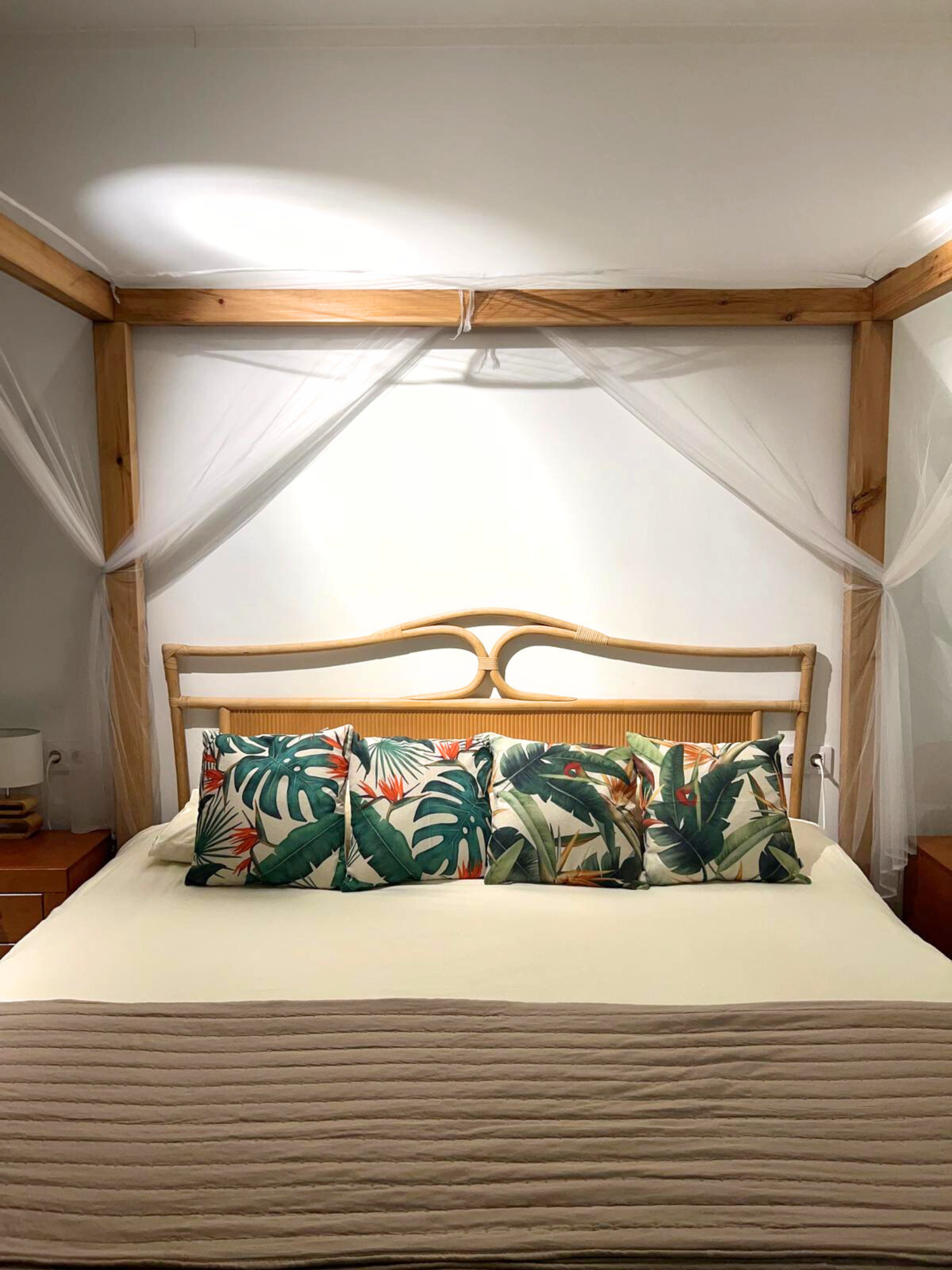 A four-poster bed with light wood frames, white netting, a tan quilt, and four pillows with tropical leaf patterns.