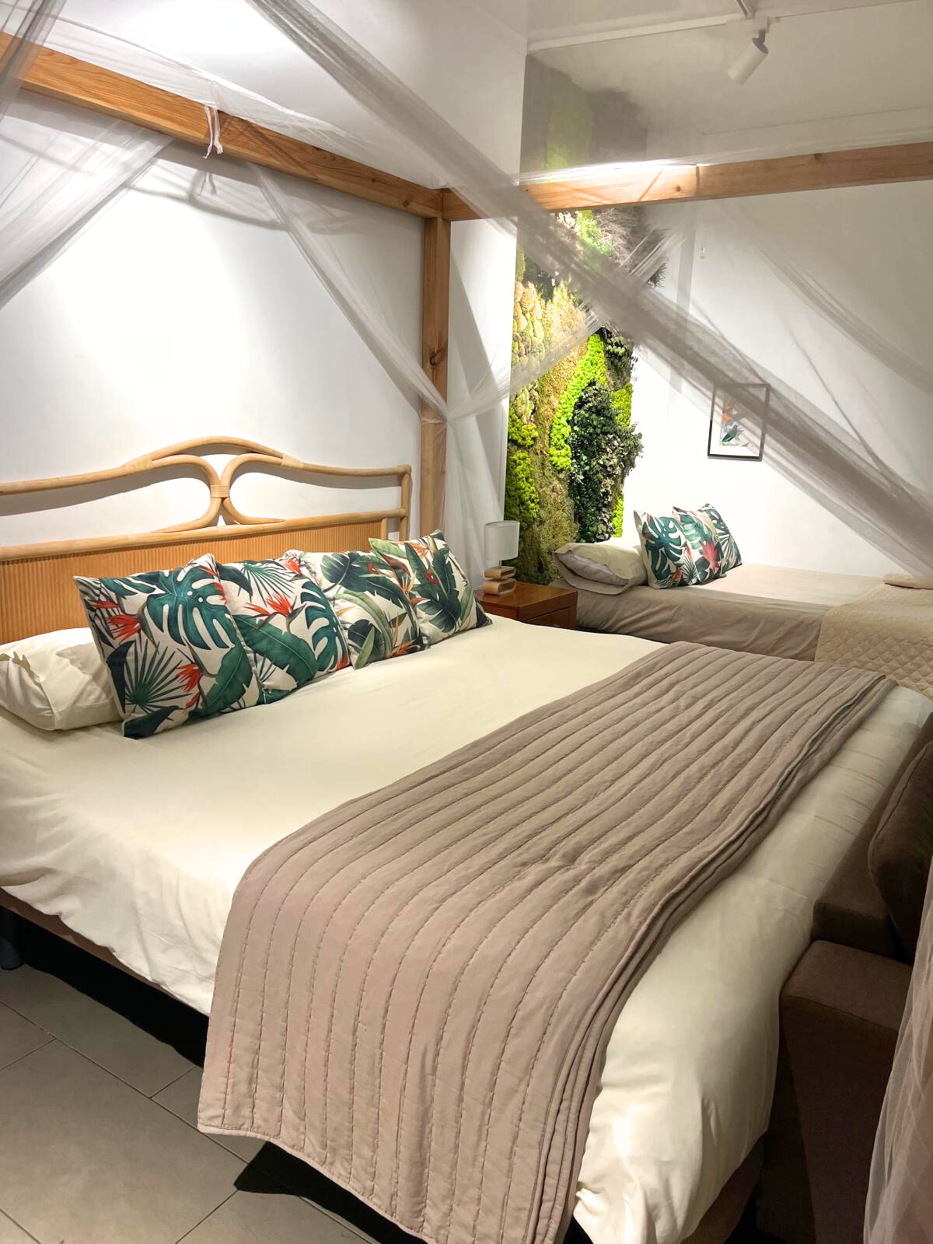 A hotel room featuring two beds with canopy frames, tropical-patterned pillows, a textured beige runner, and greenery.