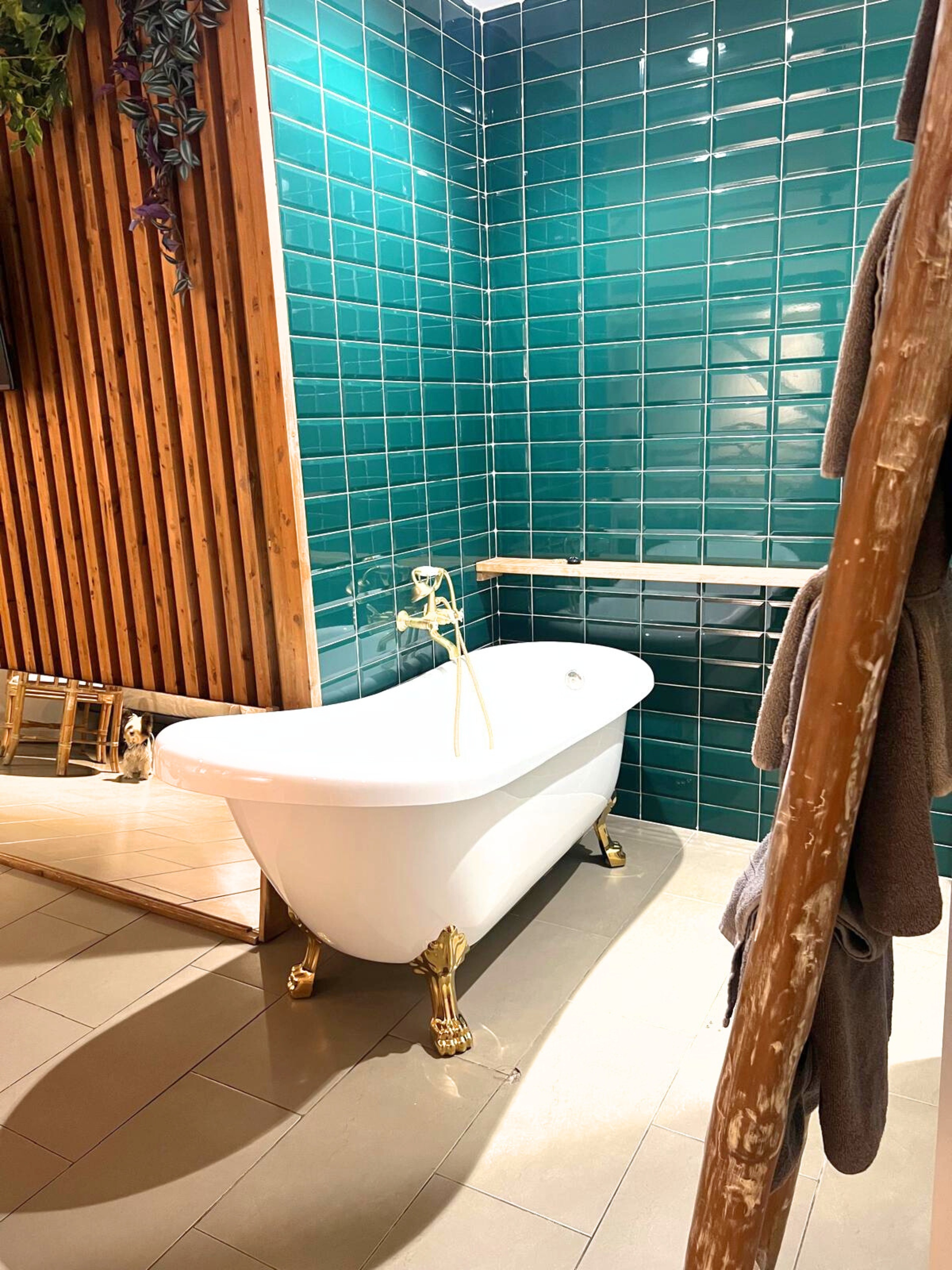 A freestanding white clawfoot bathtub with gold fixtures sits against a teal subway-tiled wall in a room with wood accents.