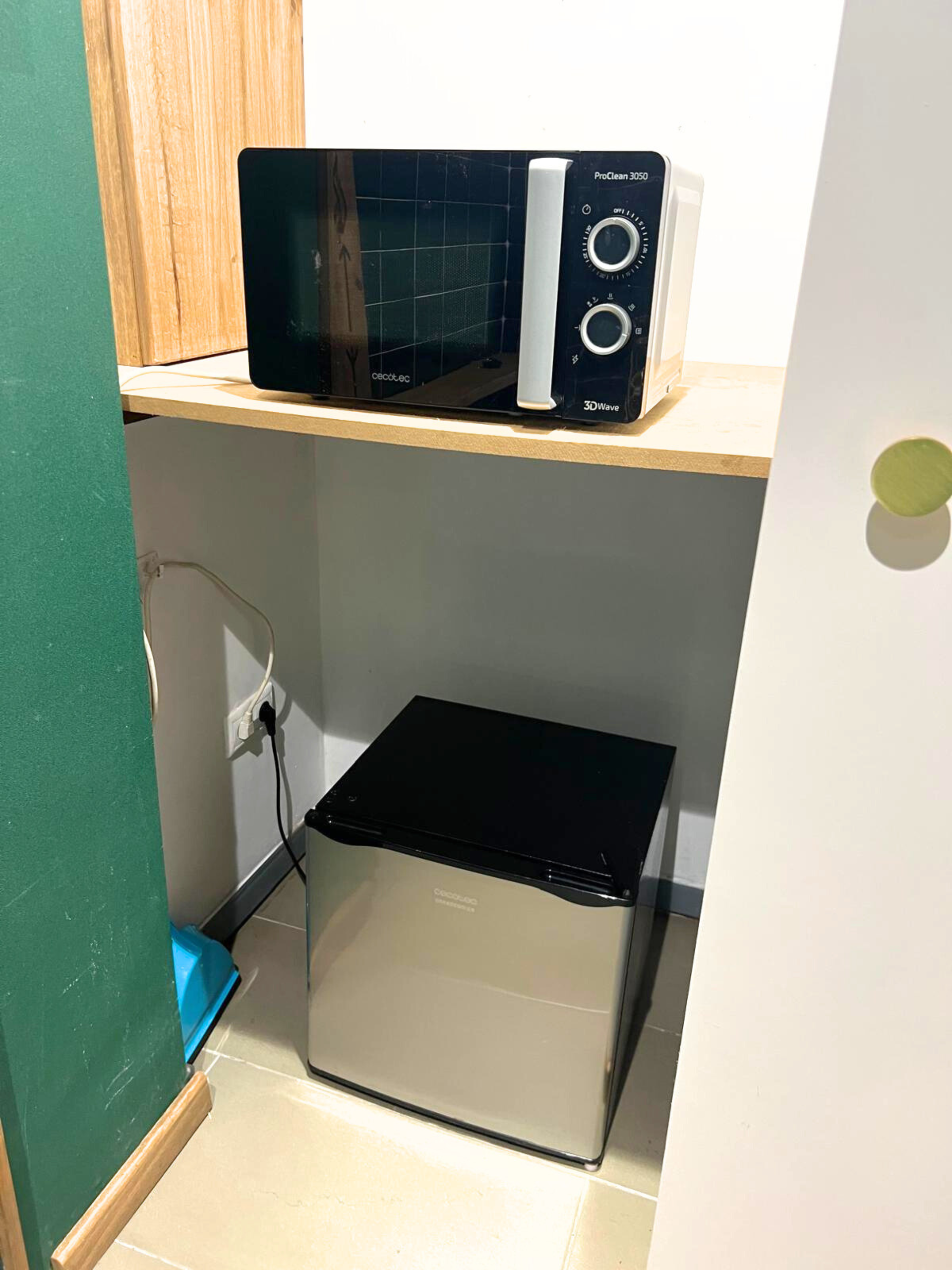 A microwave sits on a wooden shelf above a compact stainless steel refrigerator in a niche with white walls.