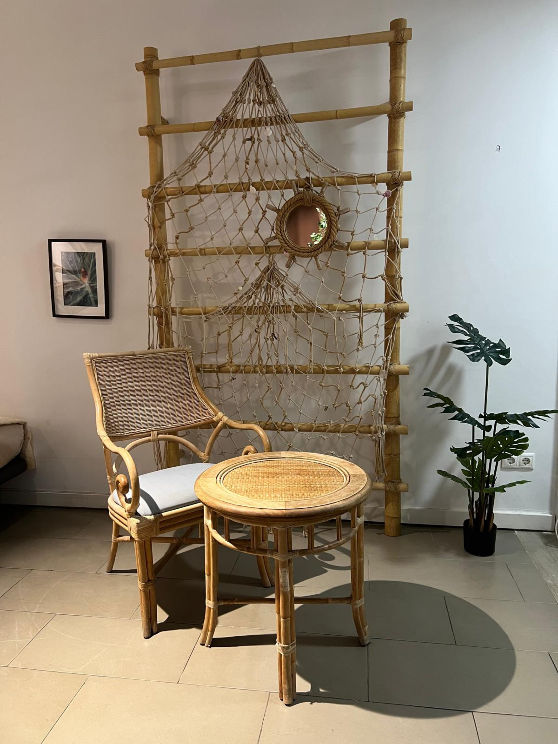 A wicker chair and round table in front of a bamboo lattice screen decorated with fishing net and a small mirror.