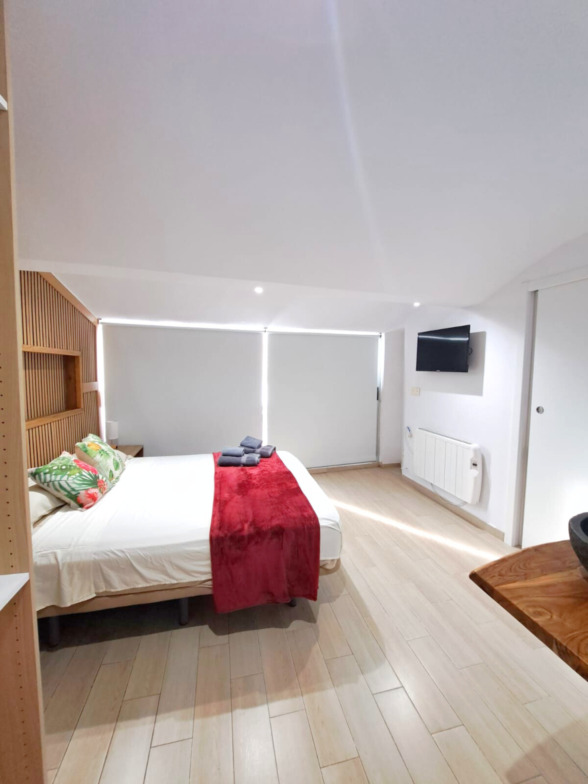 A modern bedroom with light wood flooring, a bed featuring a red throw blanket, patterned pillows, and a wall-mounted TV.