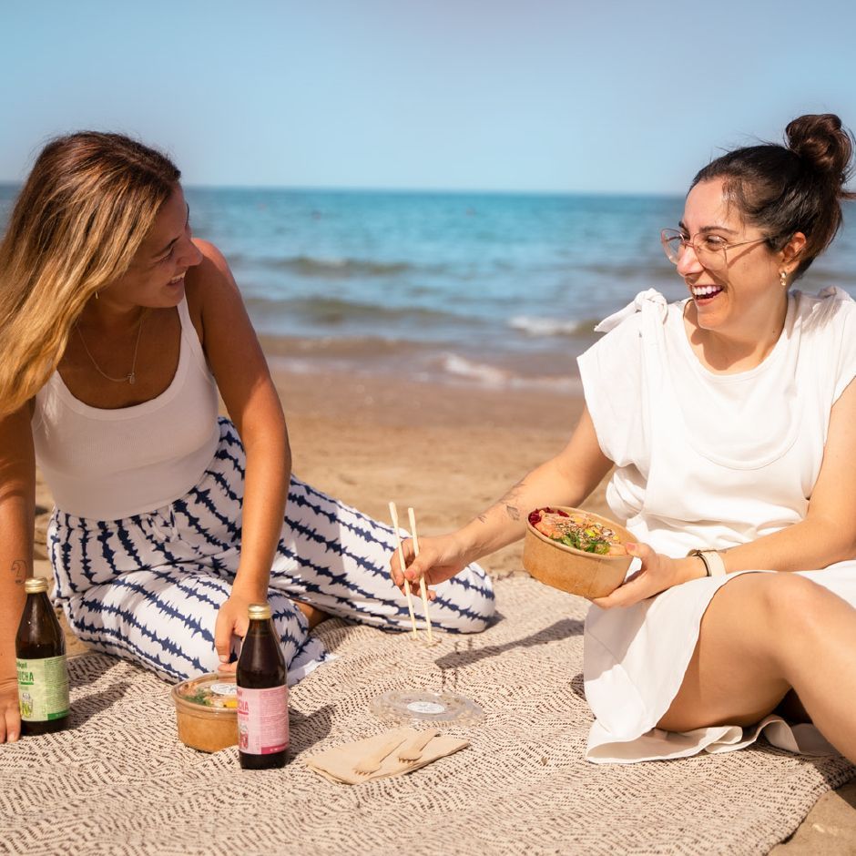 Two people enjoy a meal from bowls and bottles while sitting on a beach blanket, laughing together by the ocean.