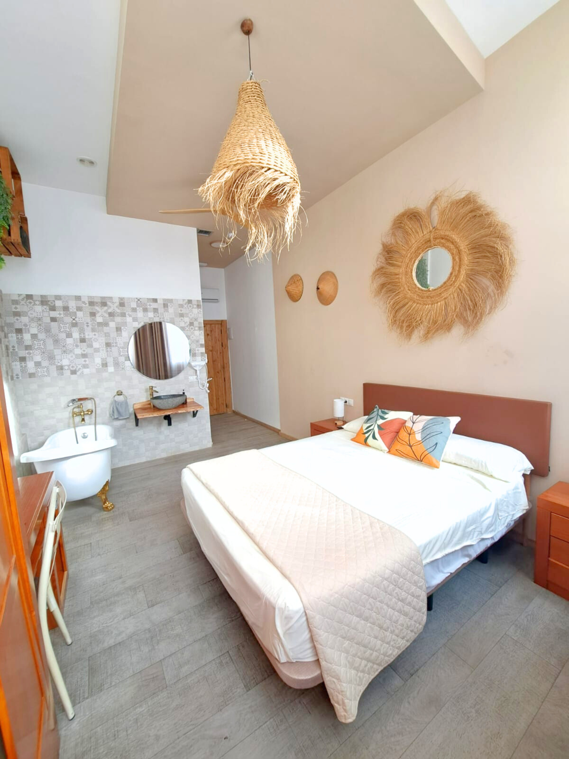 A bedroom with a queen bed, a white bathtub, and a large, textured circular mirror on a beige wall under a woven light.