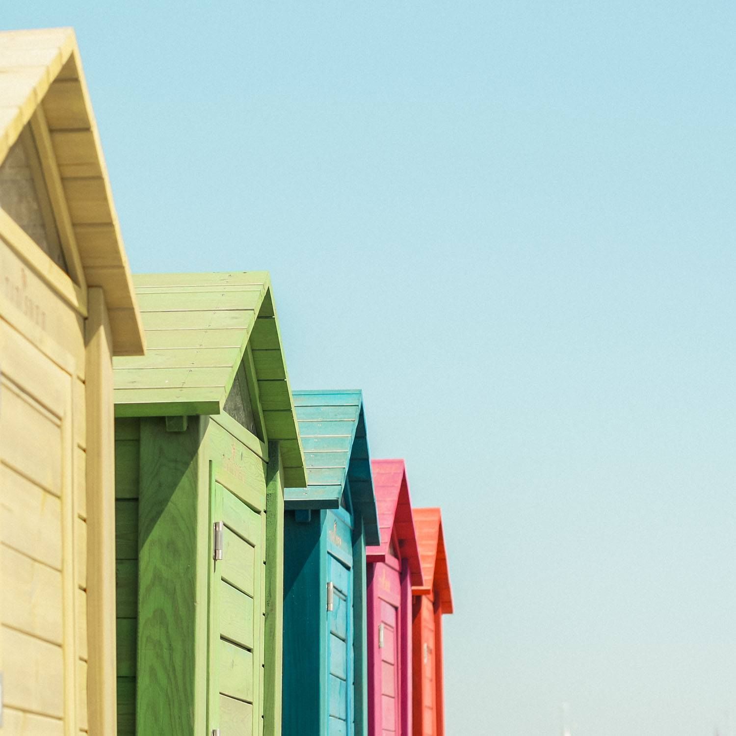 A row of colorful beach huts in yellow, green, blue, pink, and orange against a clear blue sky.