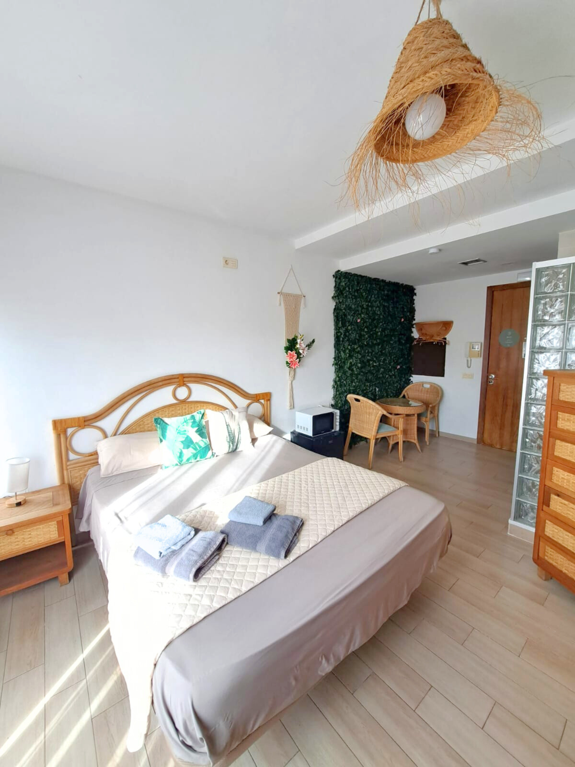 A bright bedroom featuring a queen bed with gray linens, a rattan headboard, a straw pendant light, and a faux-plant wall.