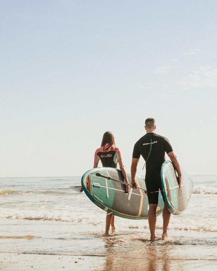 Two people walk into the ocean carrying paddleboards under their arms on a bright, sunny day.