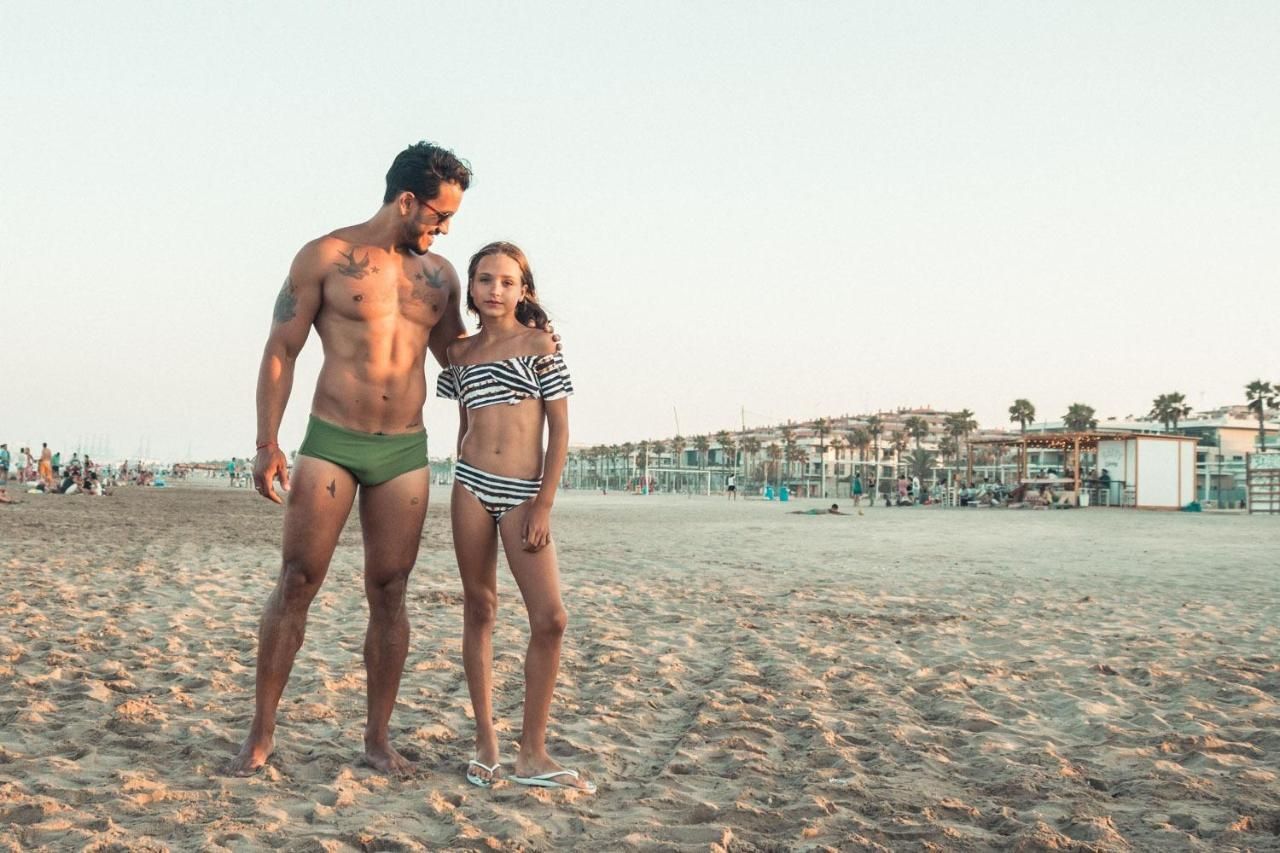 A person in green trunks and another in a striped swimsuit stand together on a sandy beach at sunset.