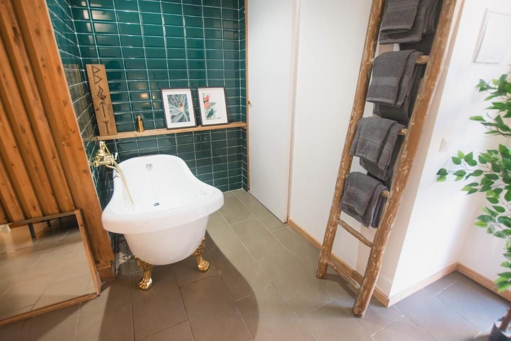 A white clawfoot bathtub sits in front of a dark green tiled wall, beside a wooden towel ladder holding grey towels.