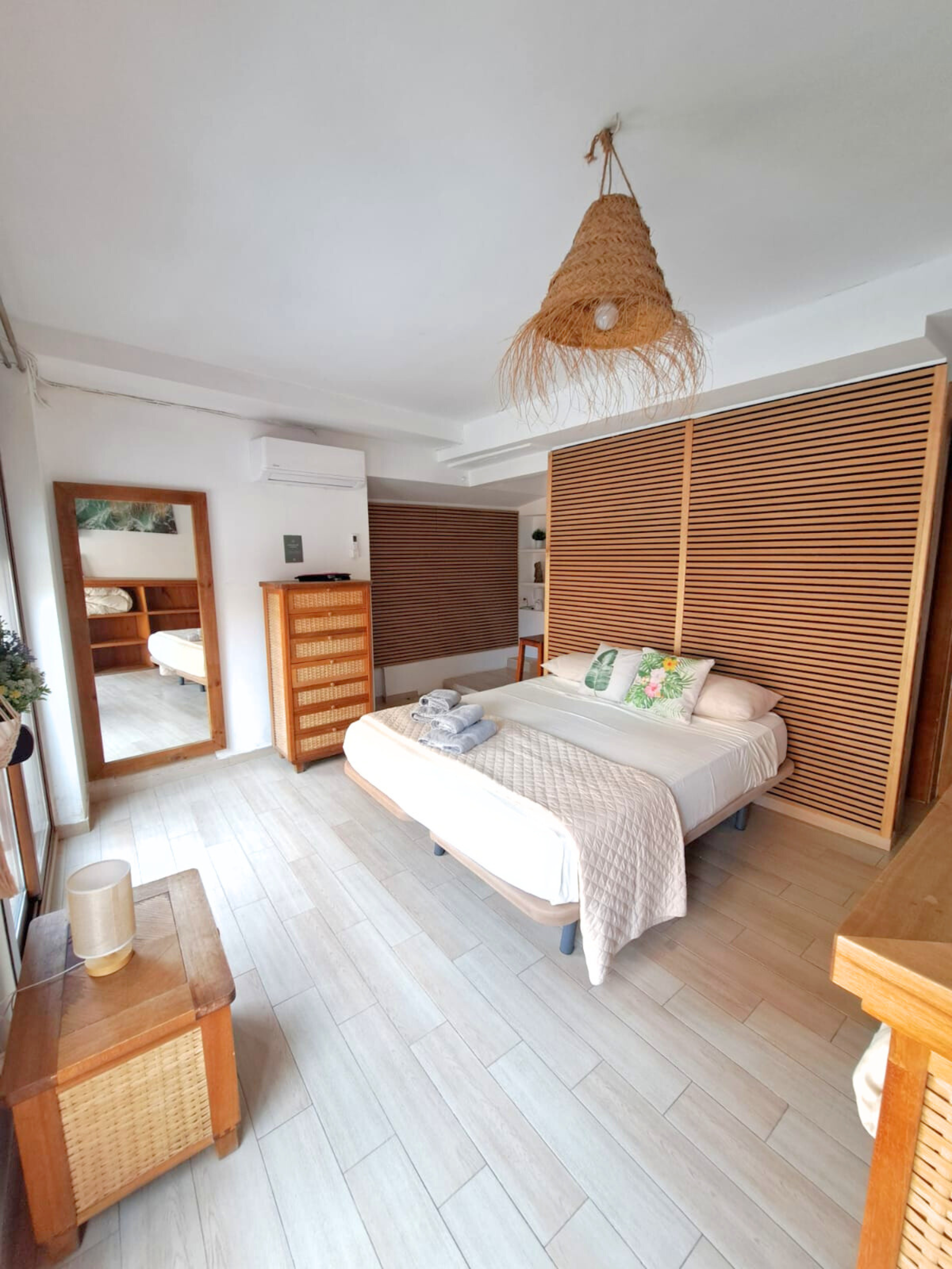 A hotel bedroom with a queen bed, a large wooden headboard, a dresser, and a floor mirror, decorated in earthy tones.