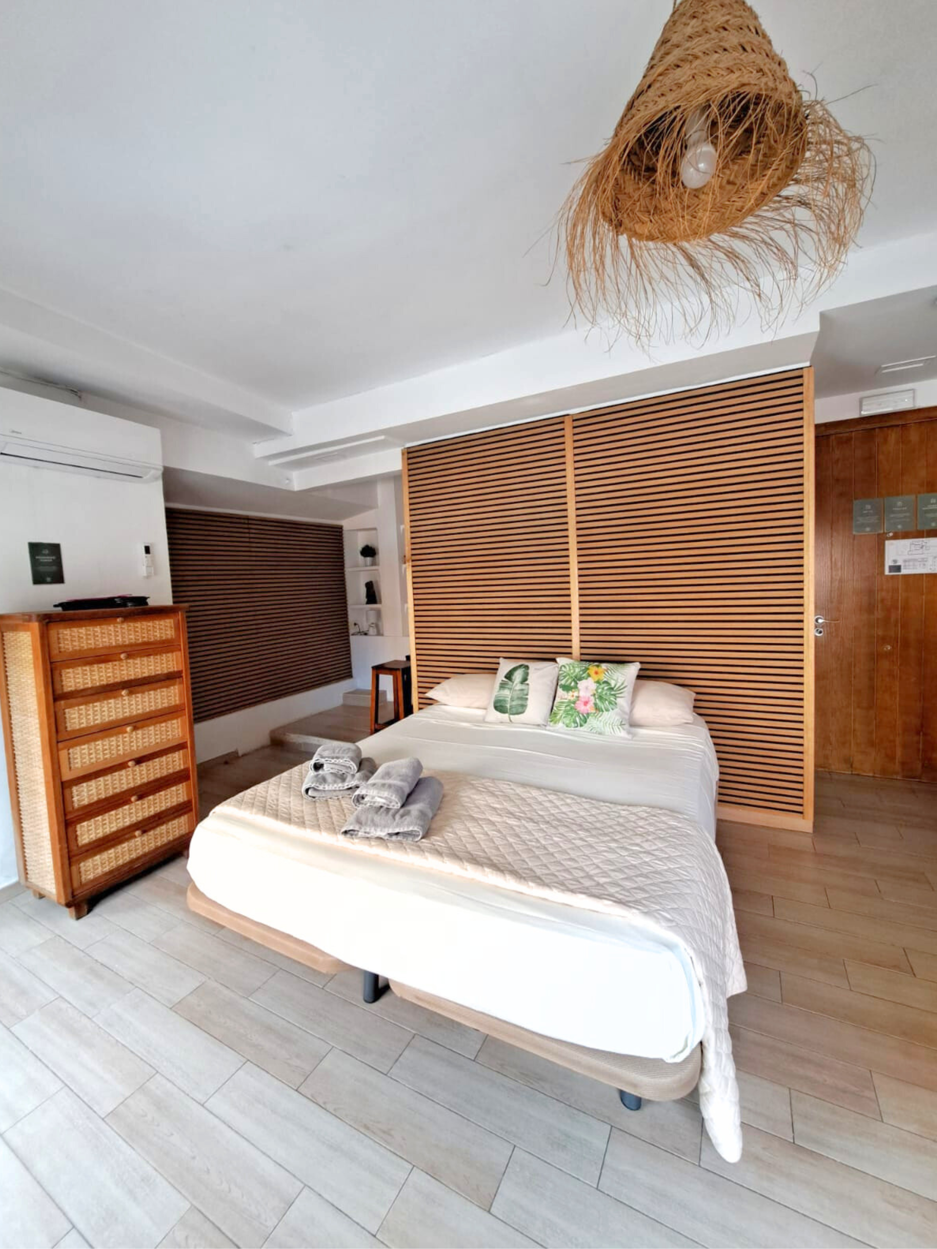A hotel room with a neatly made white bed, a wooden slatted headboard, a wicker dresser, and a woven pendant light.