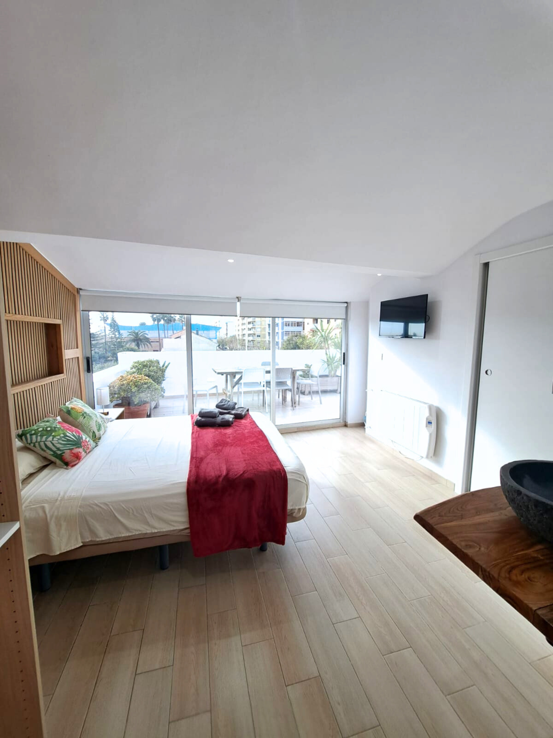 A modern hotel bedroom with a double bed featuring a red runner, wooden headboard, and a balcony view with patio furniture.