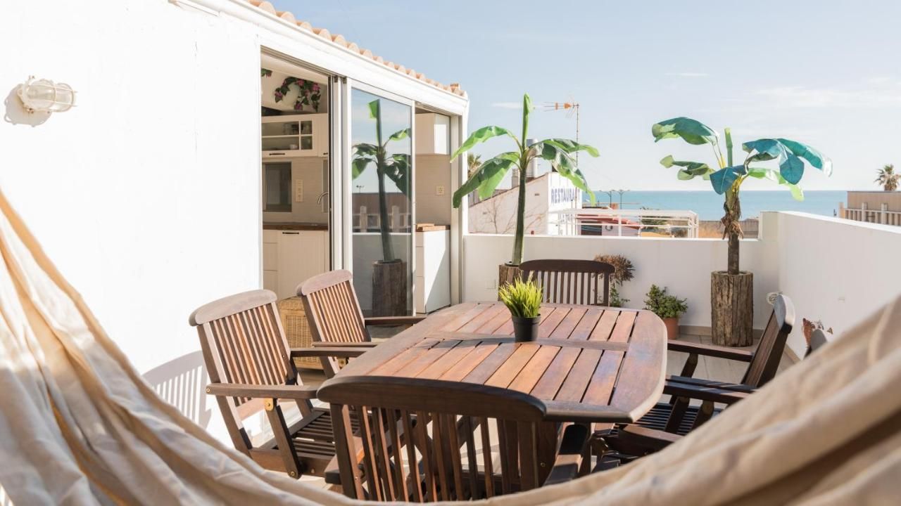 A wooden dining set on a sunlit rooftop terrace overlooking the sea, partially framed by a hammock in the foreground.