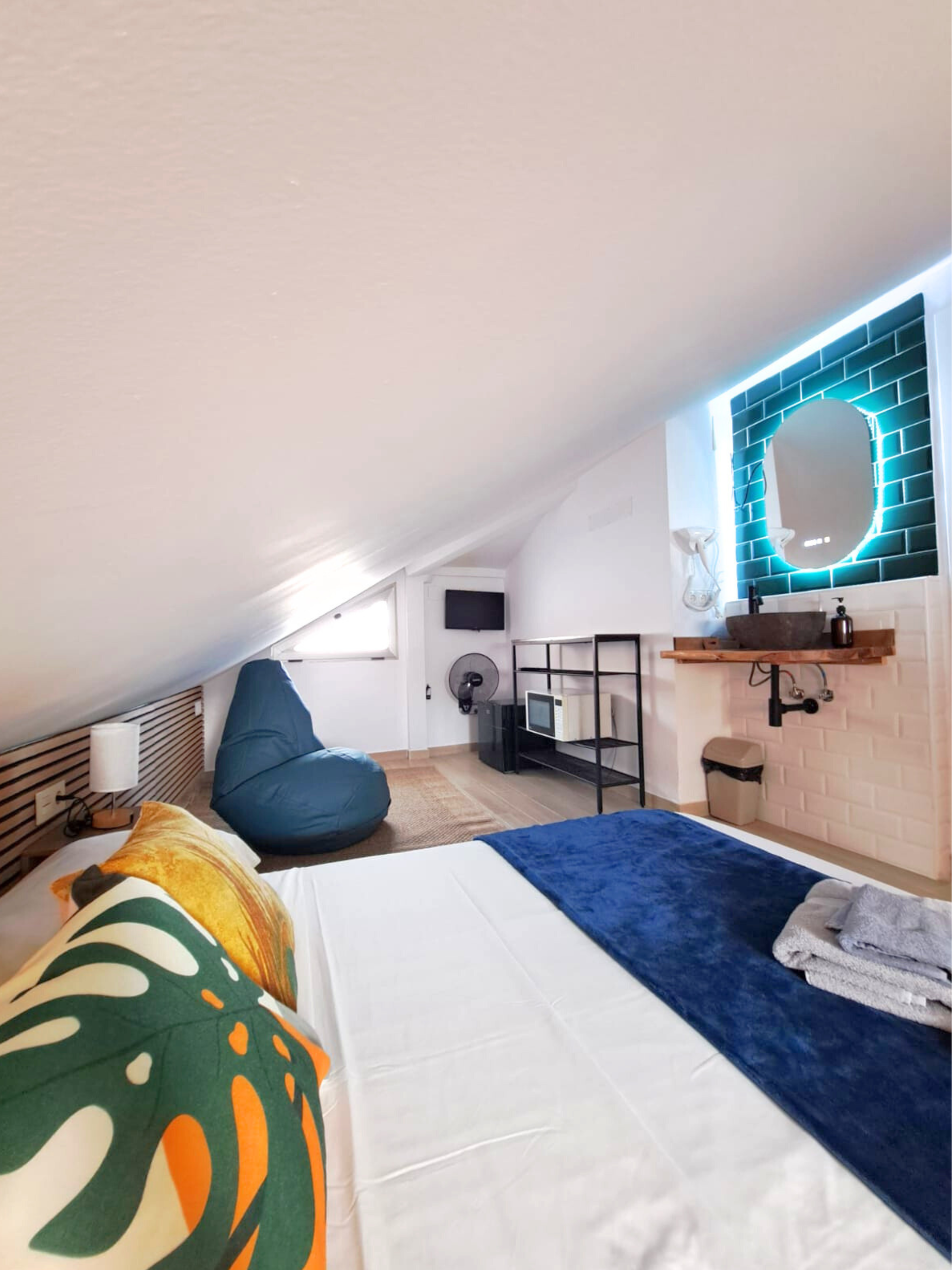 A loft bedroom with an angled ceiling, a large bed with patterned pillows, a blue beanbag chair, and a vanity sink area.