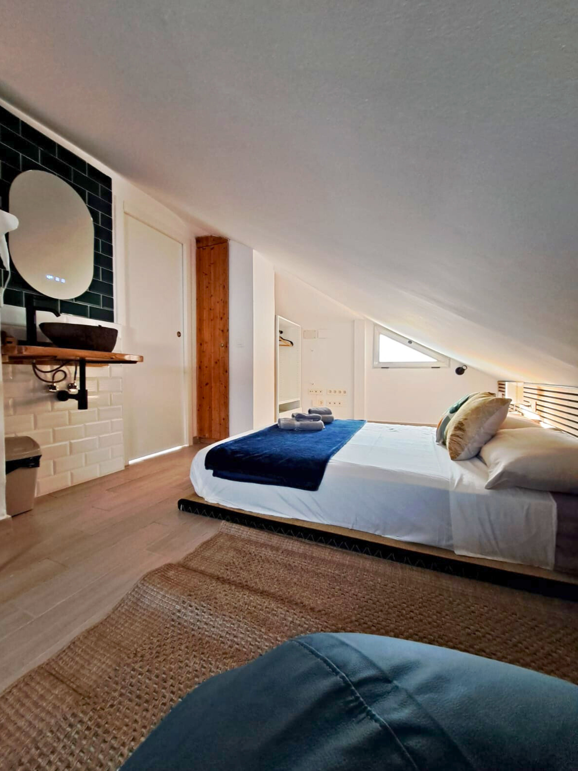 A modern attic bedroom with a low-slung bed, wooden floors, a jute rug, and a rustic sink area with a dark basin.