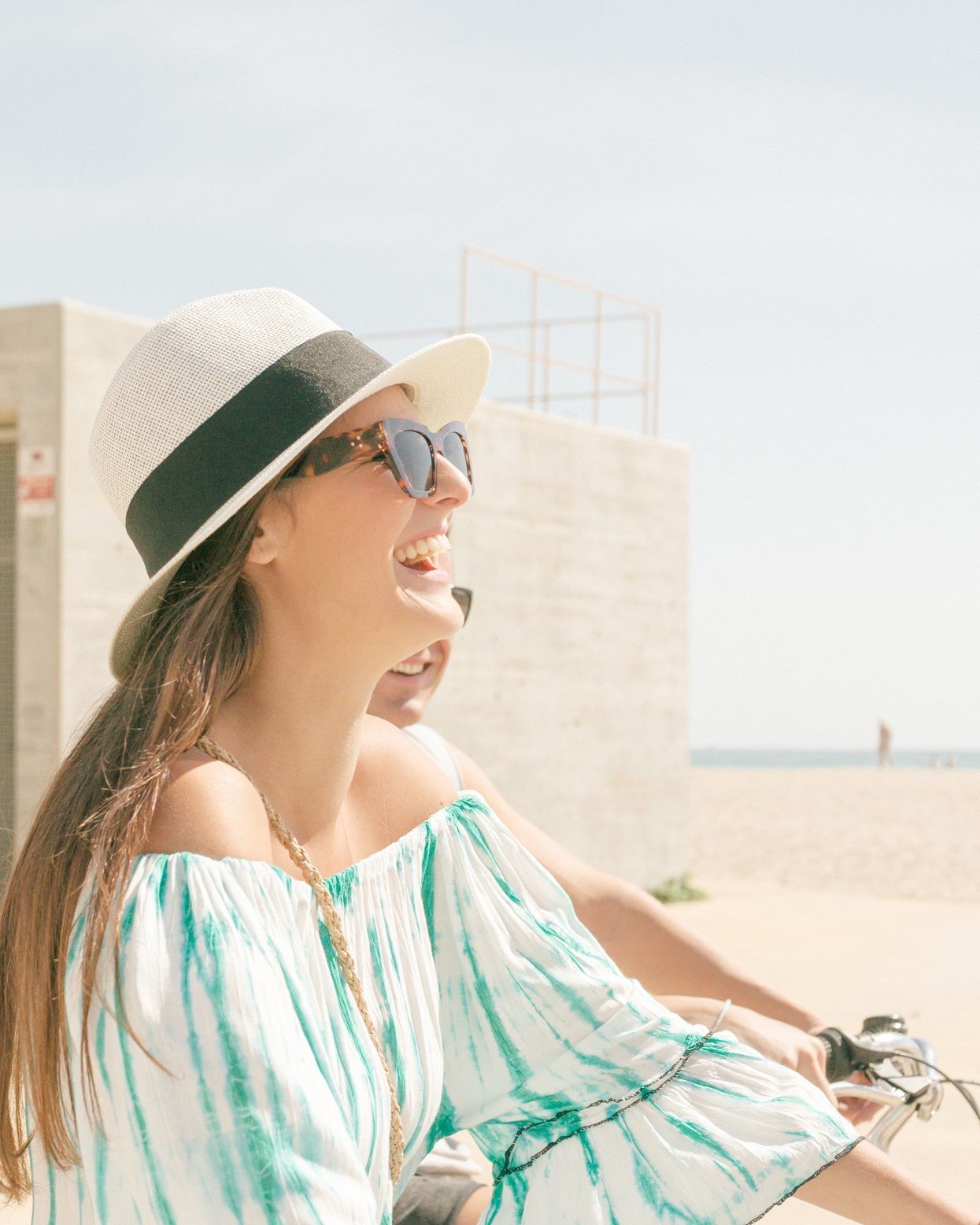 A smiling person wearing a fedora and sunglasses rides a bike outdoors near a beach building.