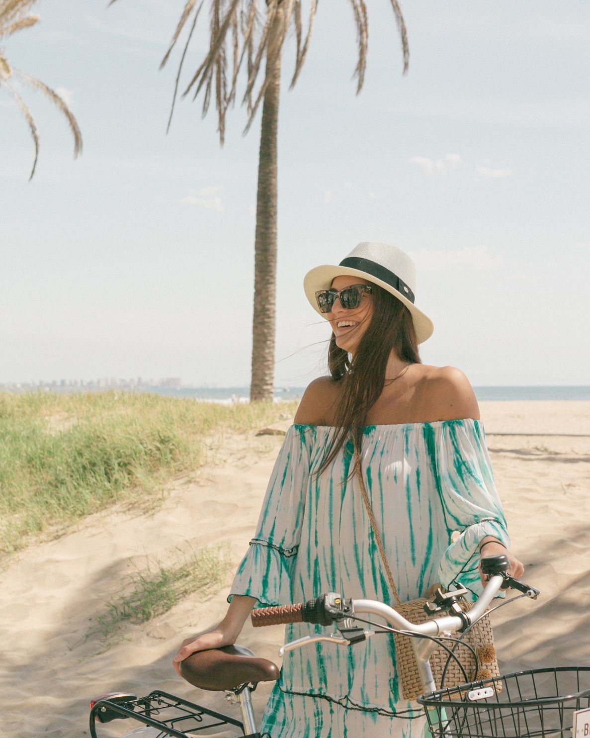 A person in a sun hat and patterned dress smiles while standing with a bicycle on a sandy beach under palm trees.