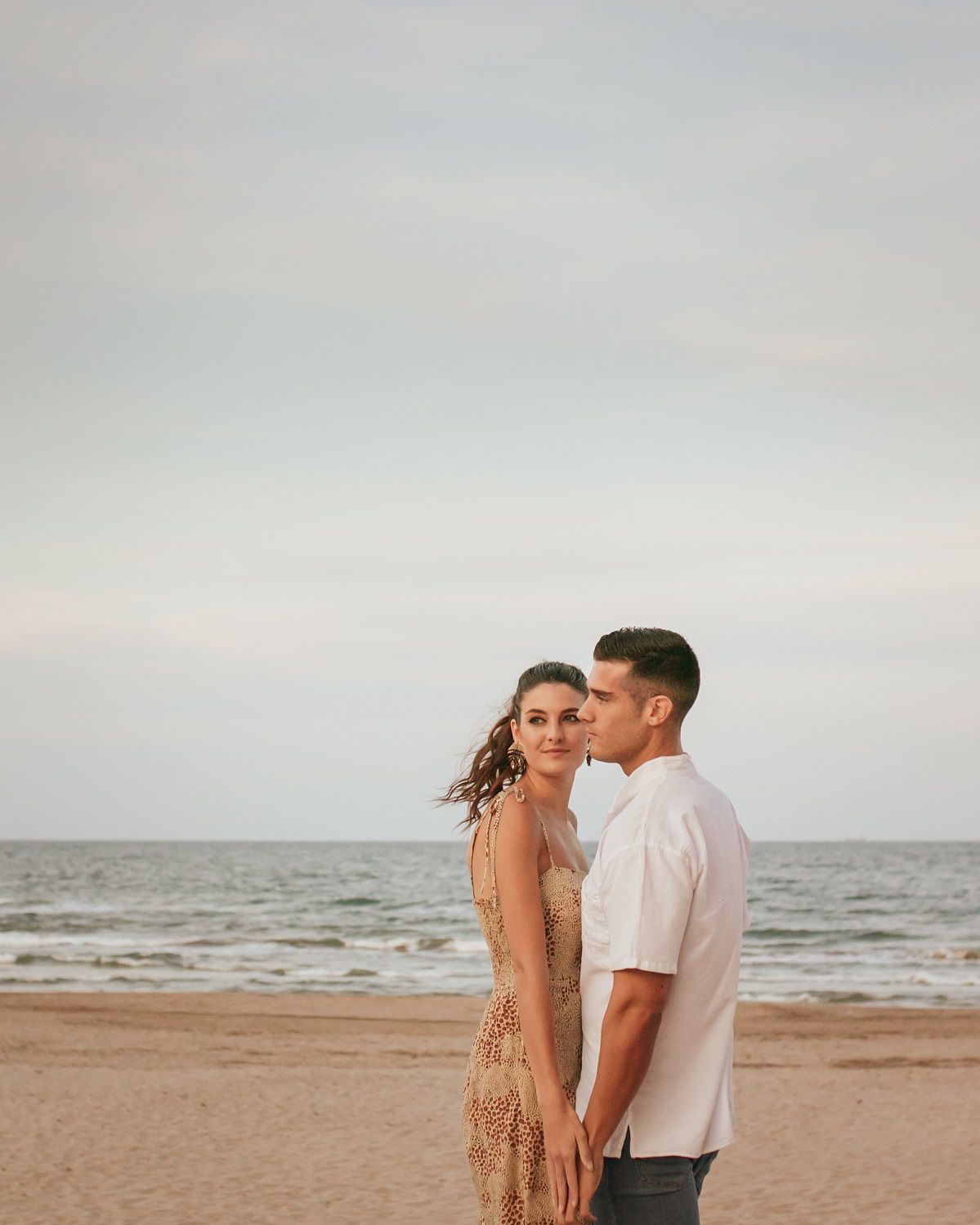 A couple holds hands while standing on a sandy beach, gazing out toward the ocean under a soft, overcast sky.