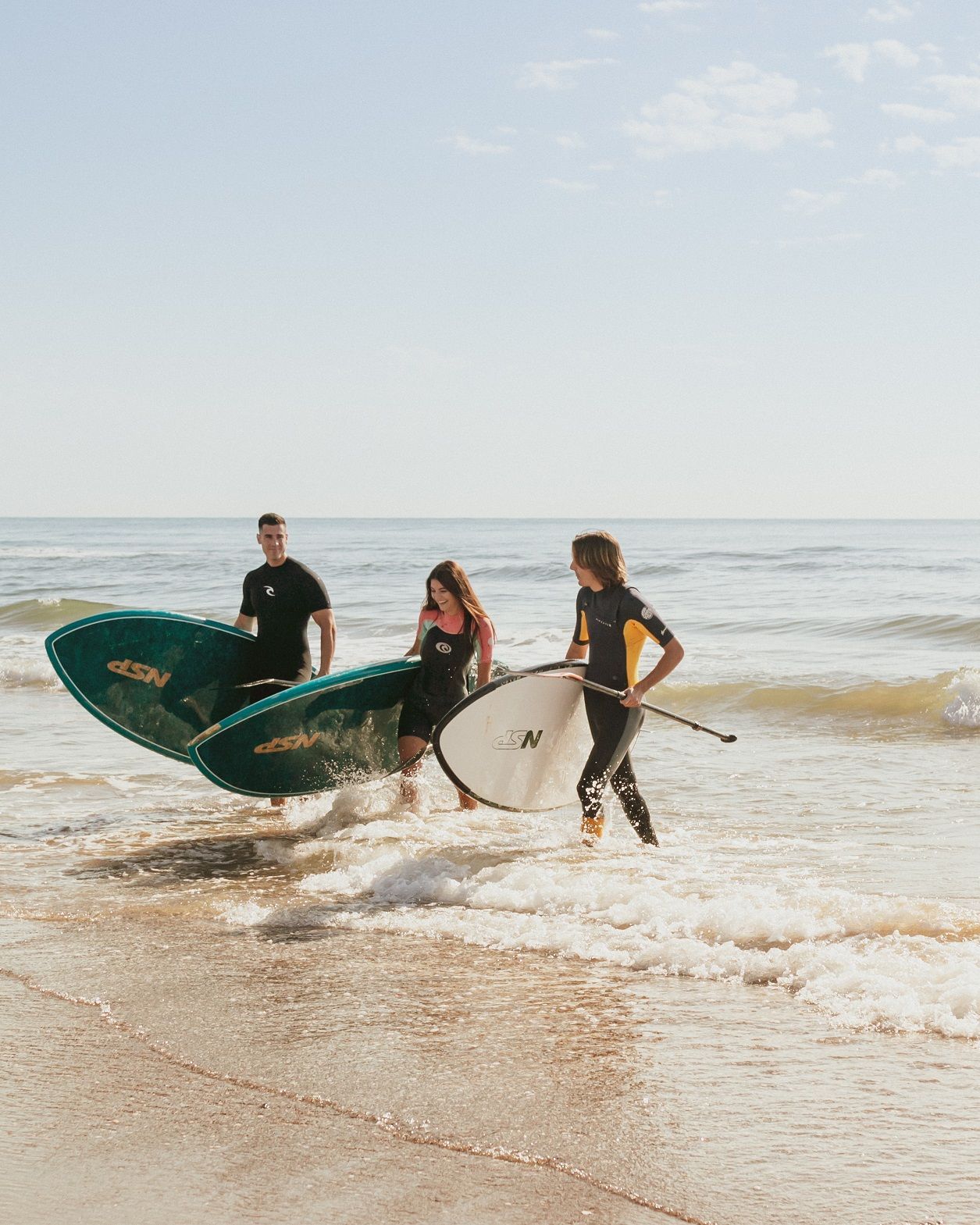 Three people in wetsuits carry stand-up paddleboards into the shallow water at the beach on a sunny day.