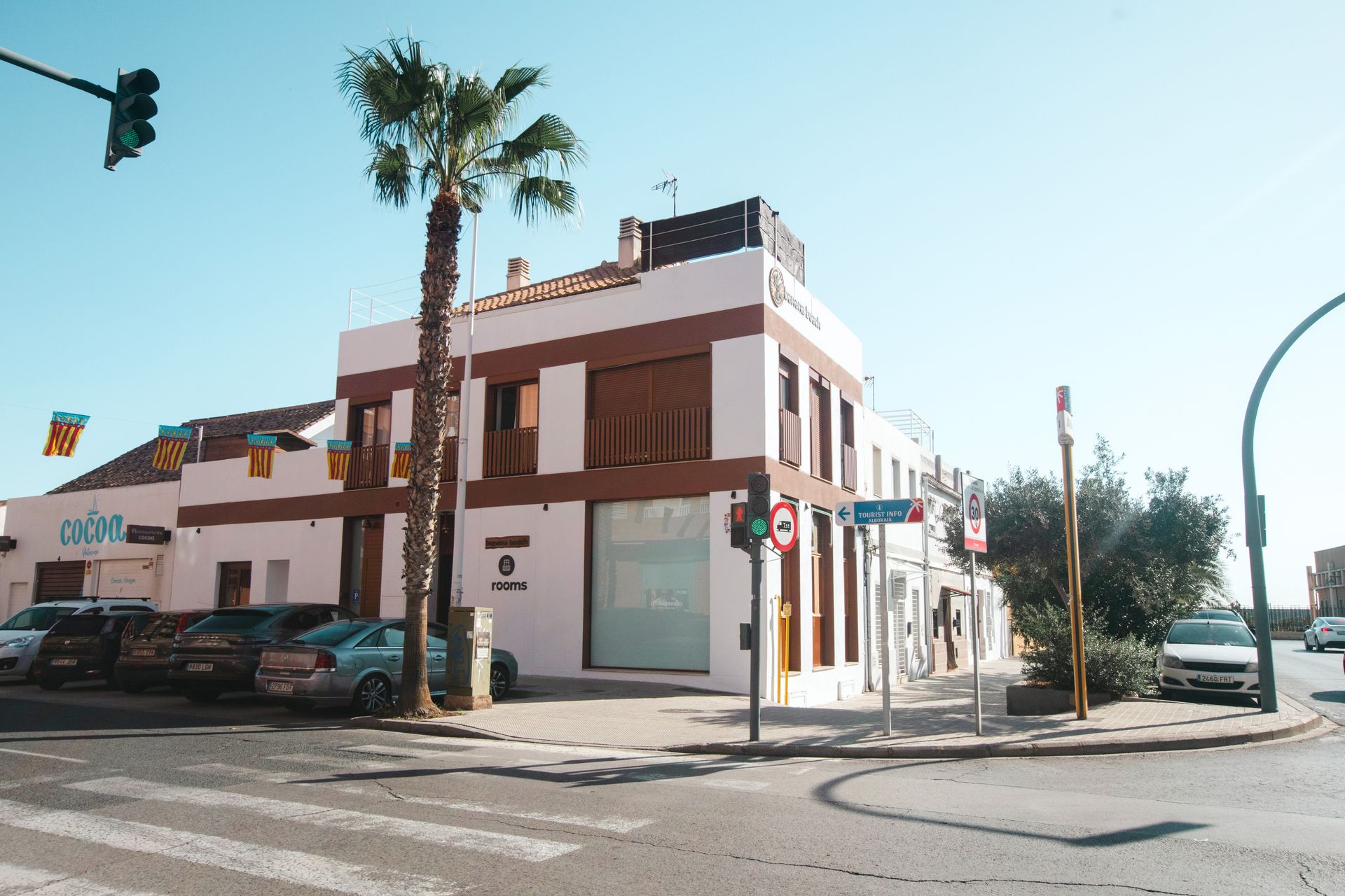 A white, two-story commercial building with brown trim and a palm tree on a sunny corner intersection with parked cars.