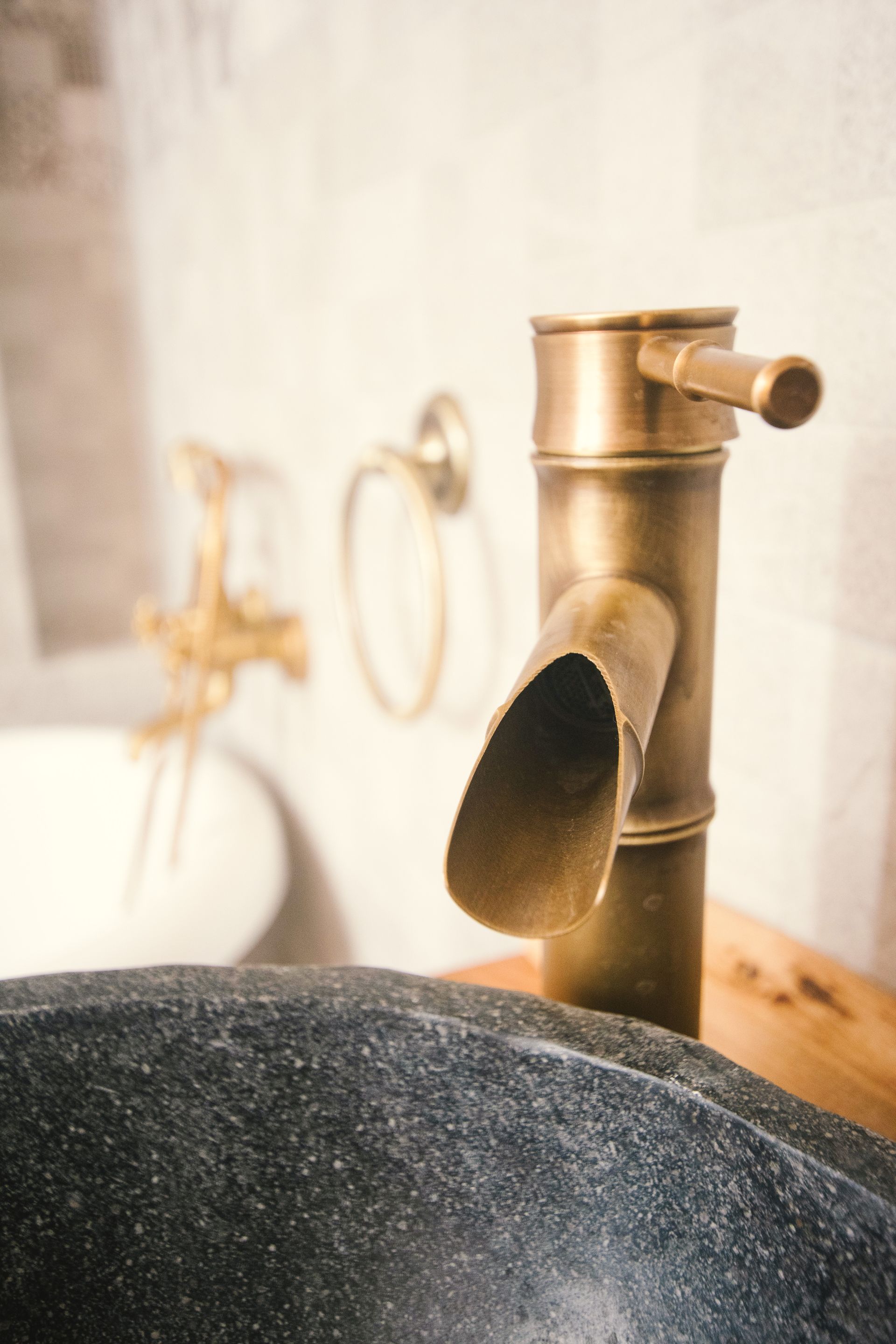 A rustic brass faucet with a slanted spout stands behind a dark, textured stone vessel sink in a tiled bathroom.