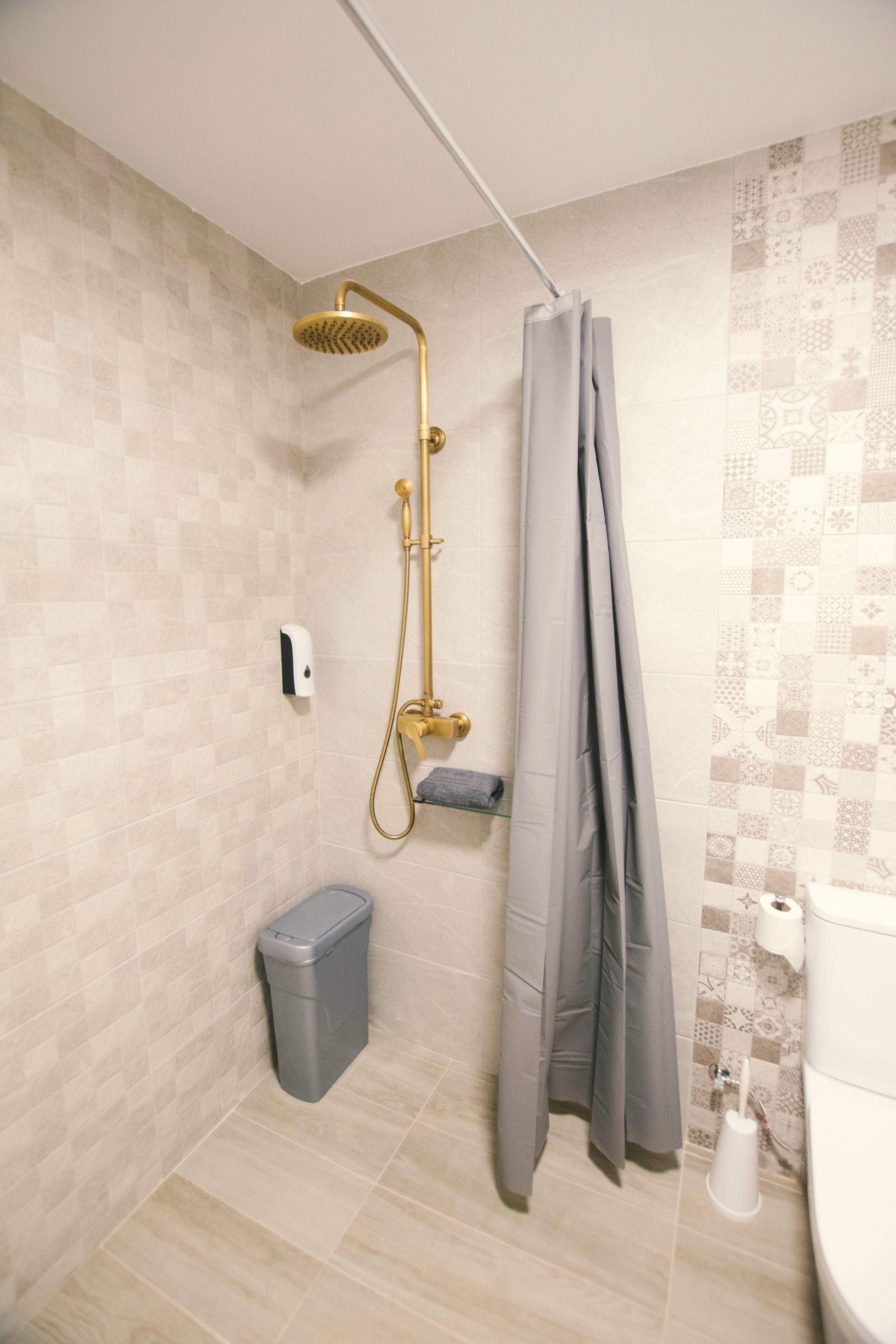 A modern bathroom with a gold shower fixture, grey shower curtain, and a grey trash bin against light tiled walls.
