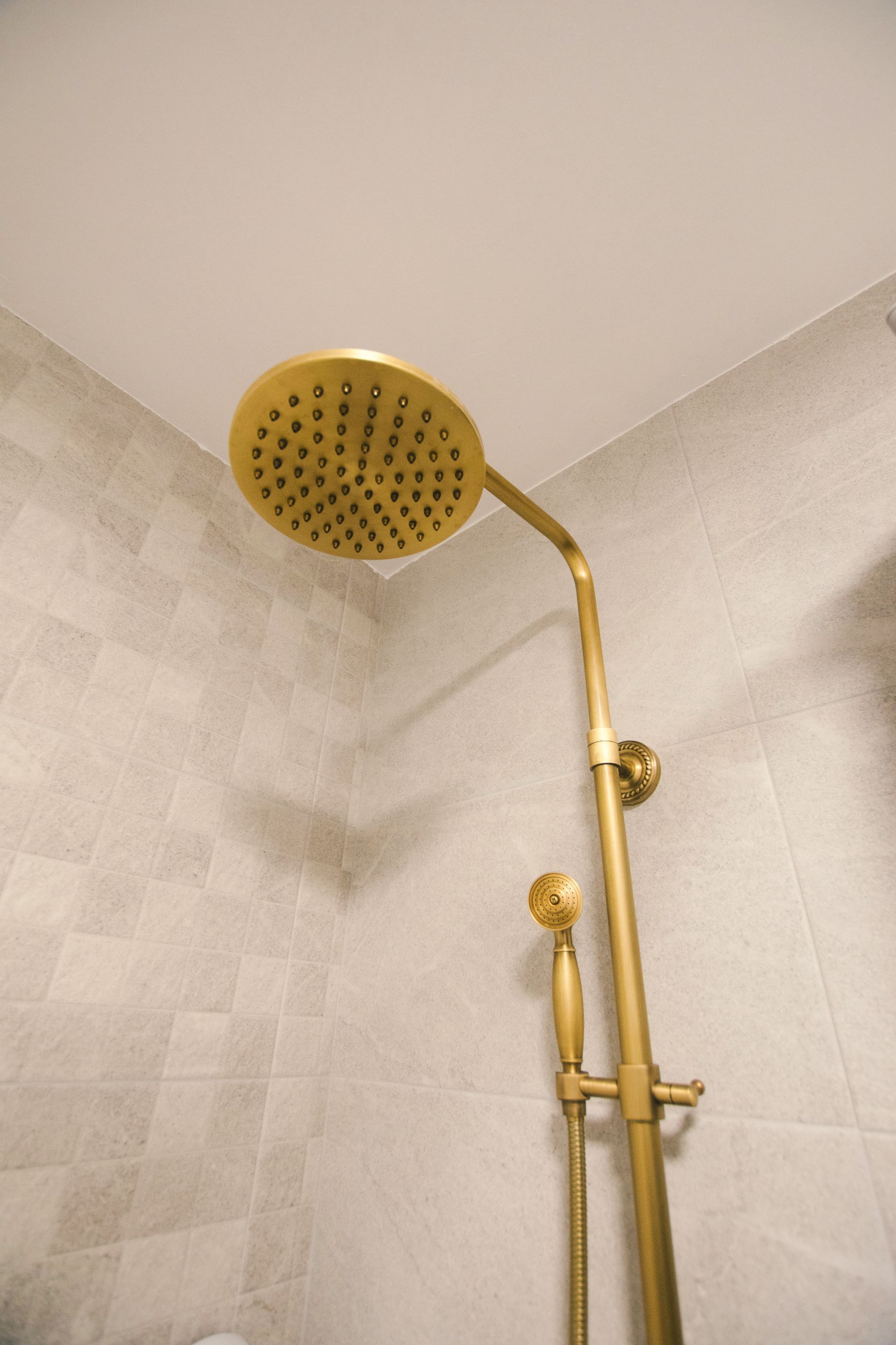 A gold-toned shower fixture with a round rain showerhead and a handheld attachment mounted against light tiled walls.