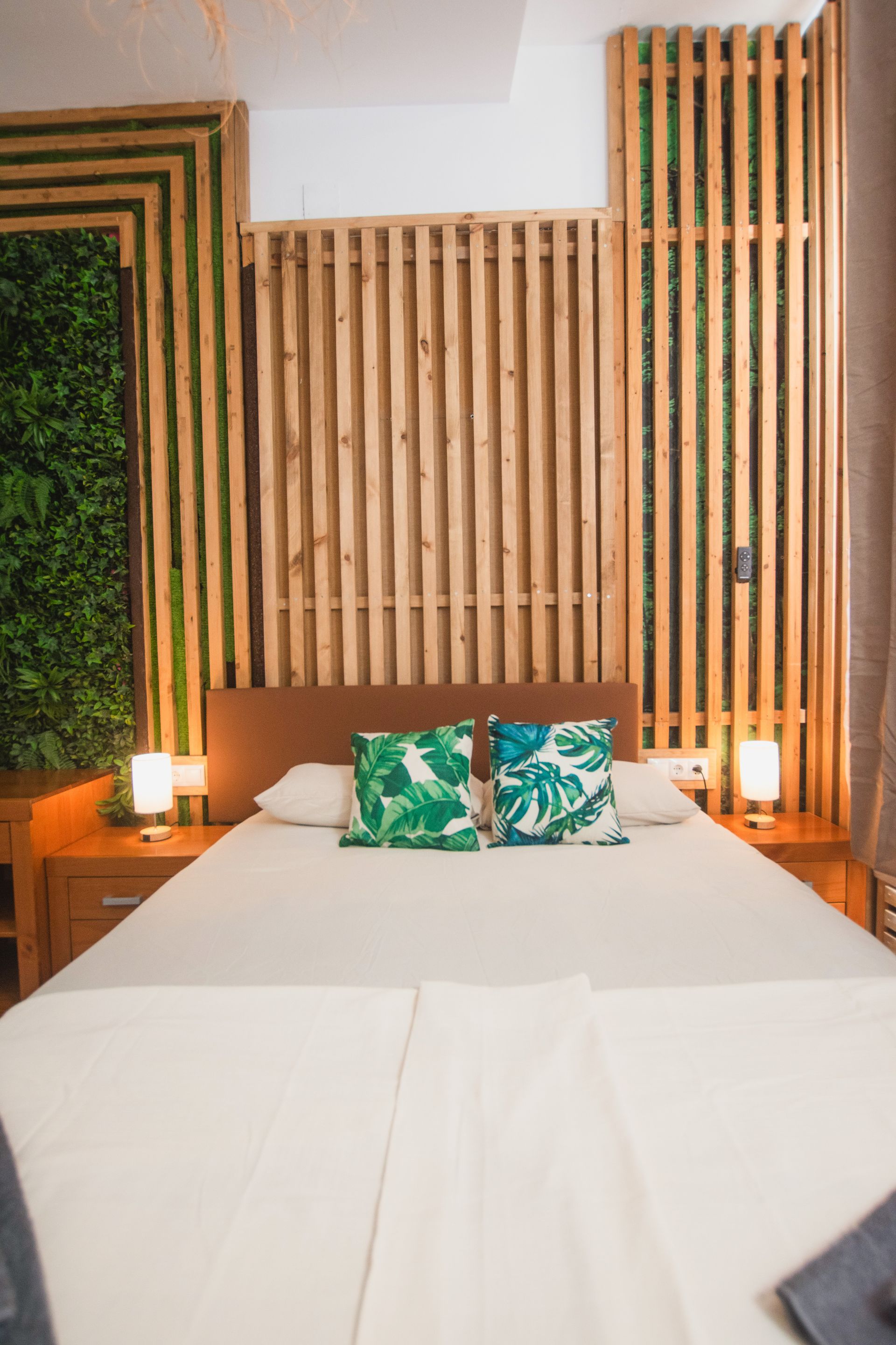 A hotel bedroom with a bed, two green tropical-patterned pillows, light wood wall paneling, greenery, and two lamps.