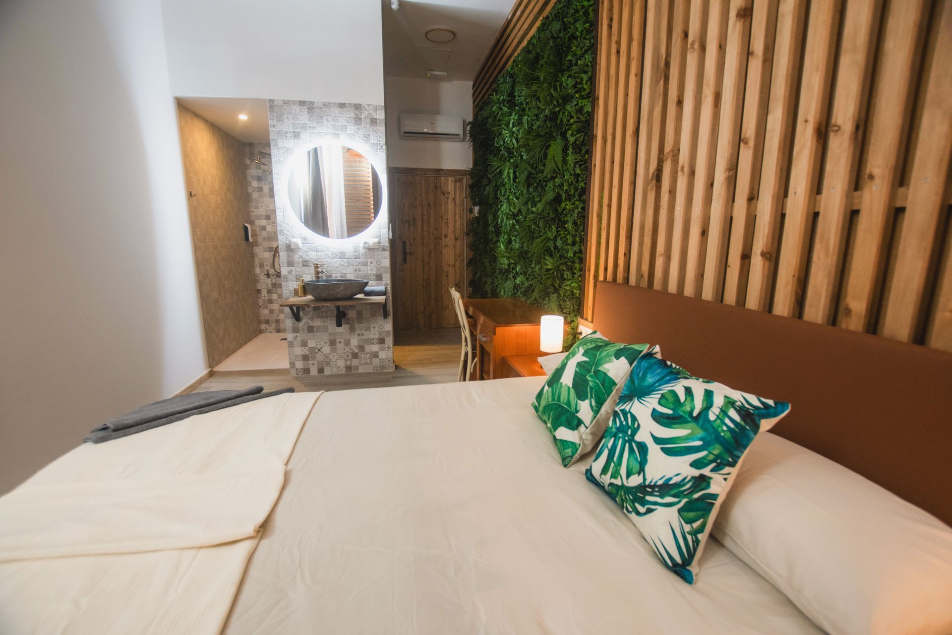 A modern hotel room with a bed featuring tropical print pillows, a wooden slat headboard, and an open-concept bathroom.
