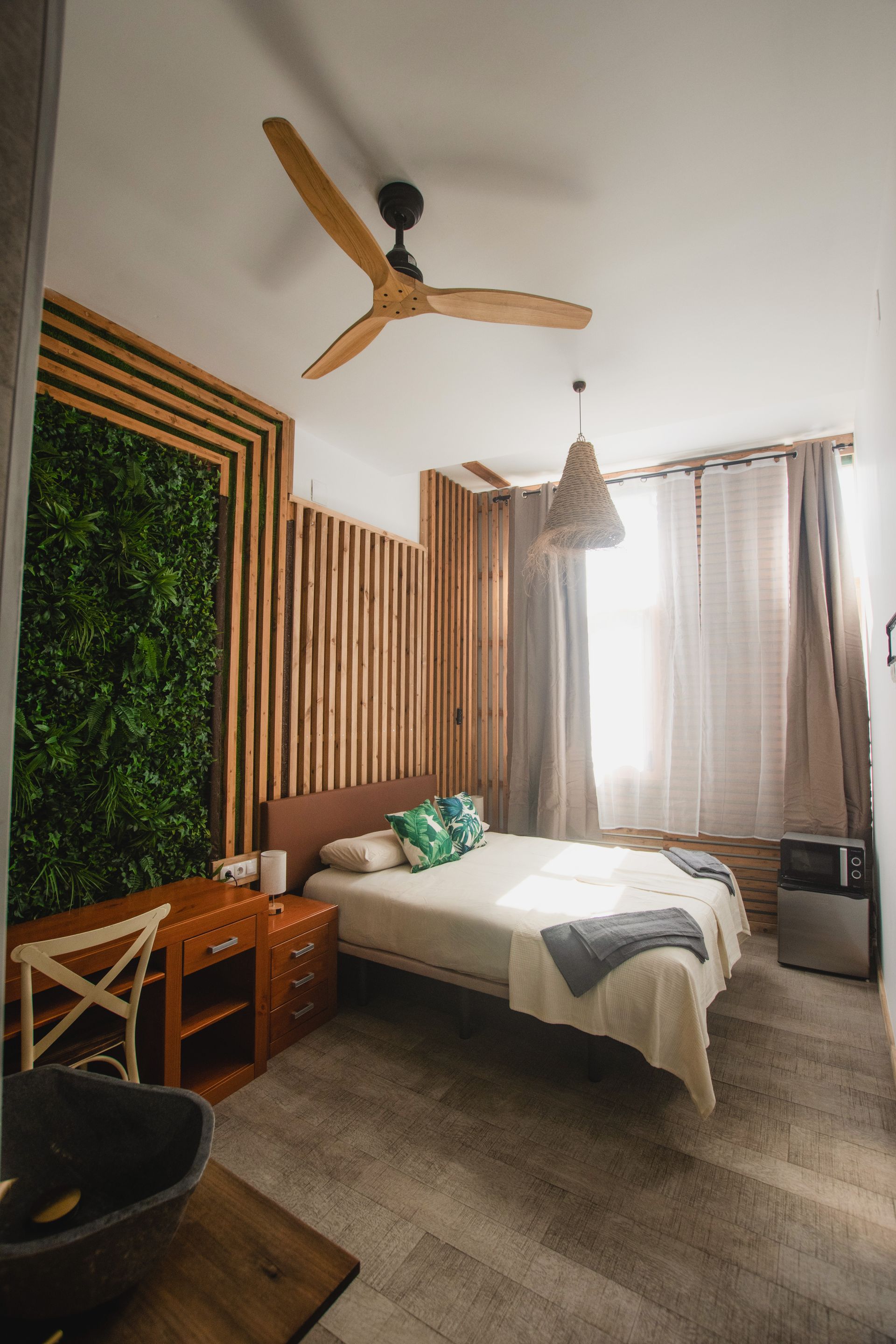 A modern bedroom features a wooden slat accent wall with greenery, a bed with light linens, a ceiling fan, and curtains.