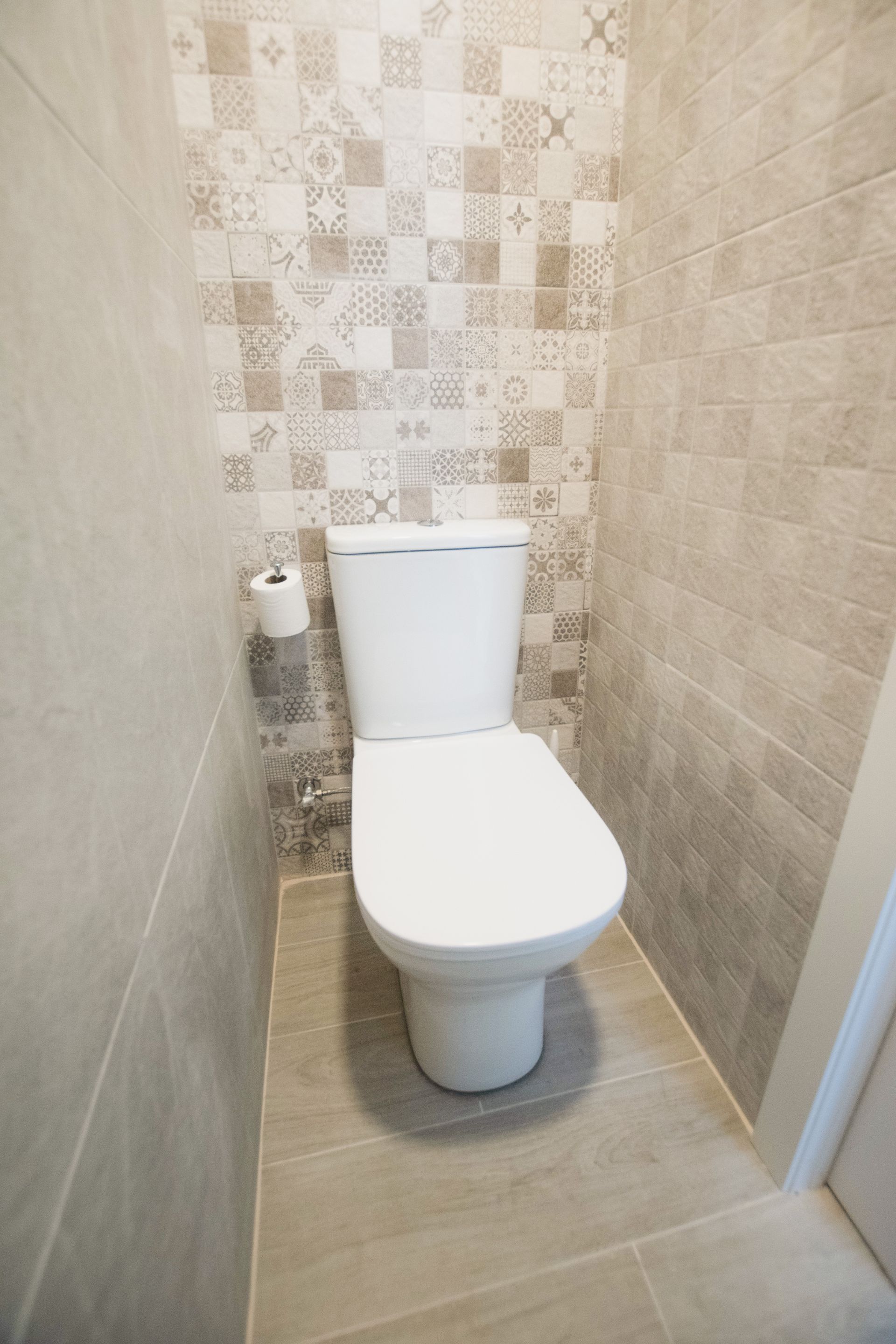 A white toilet centered in a small bathroom with patterned back tiles and neutral stone-look floor and side wall tiles.