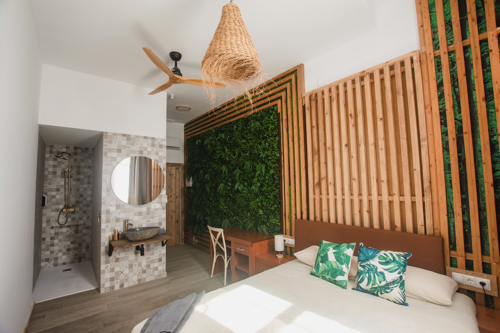 A bedroom with a bed, a wooden headboard, a green plant wall, a ceiling fan, and an open-concept bathroom vanity.