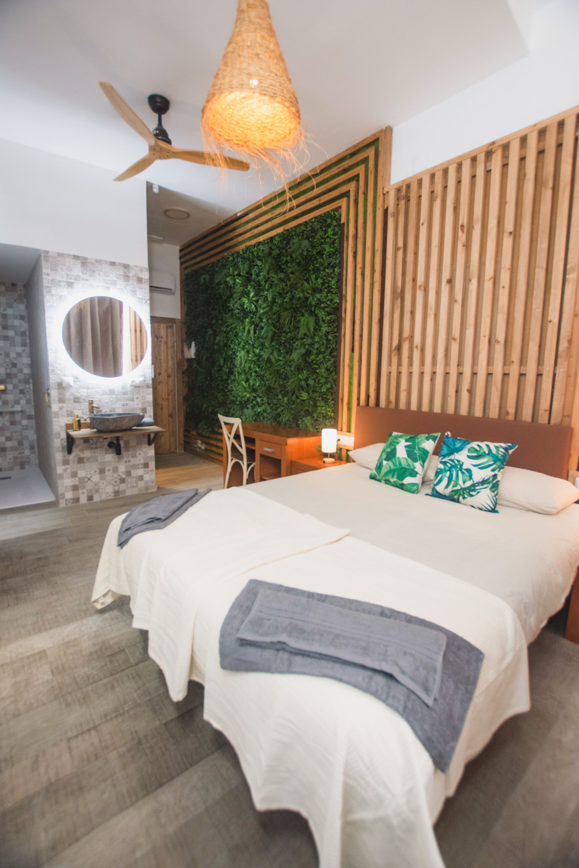 A modern bedroom featuring a bed with white linens, a living plant wall, a wooden slatted headboard, and a wicker lamp.