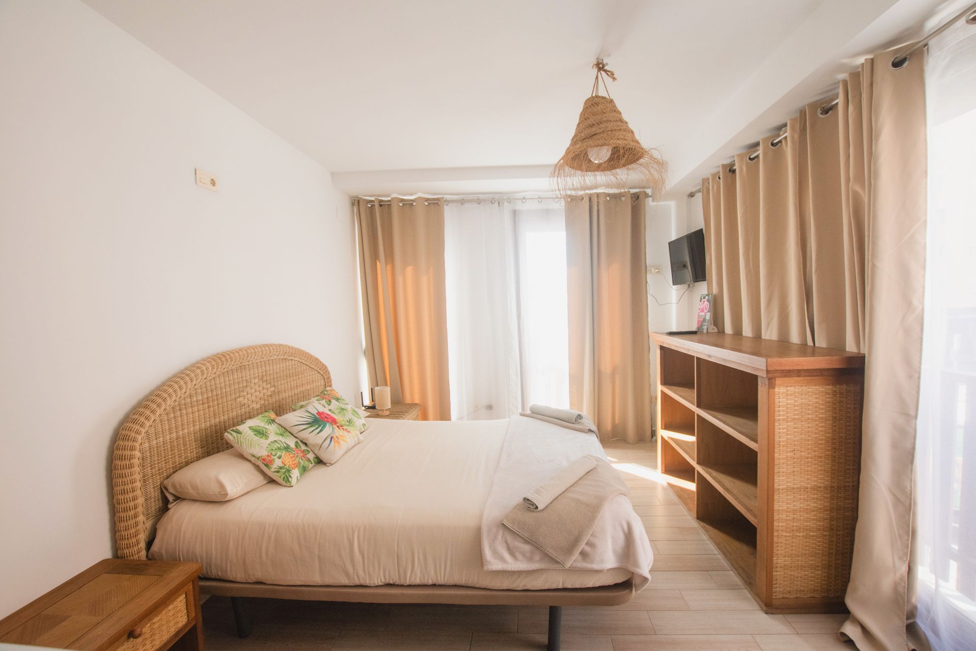 A bright bedroom with a natural wicker headboard, beige bedding, matching curtains, and a rattan pendant light.
