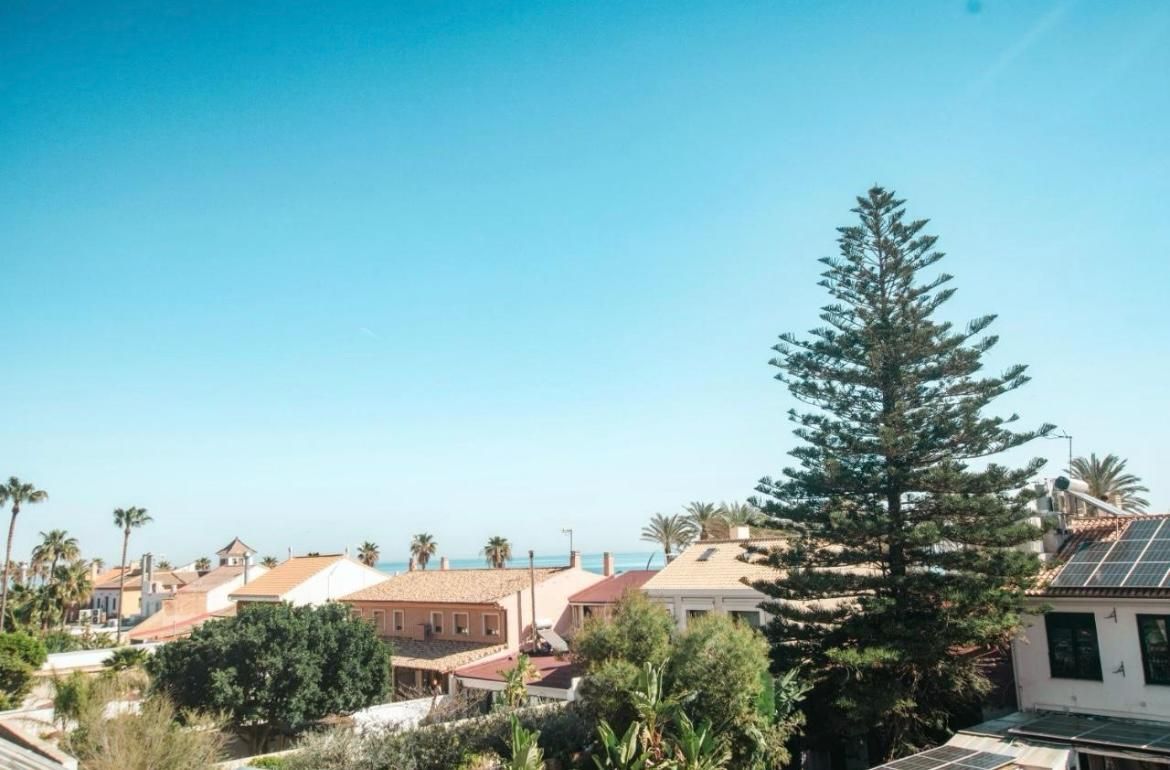 A wide-angle view of houses with terracotta roofs under a clear blue sky, featuring a tall evergreen tree on the right.