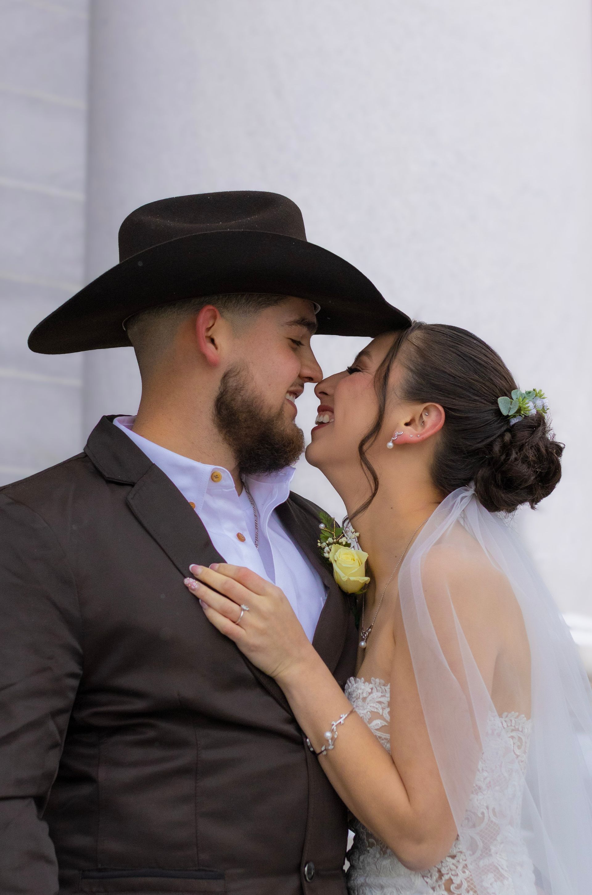 A bride and groom are kissing in front of a white wall . the bride is wearing a cowboy hat.