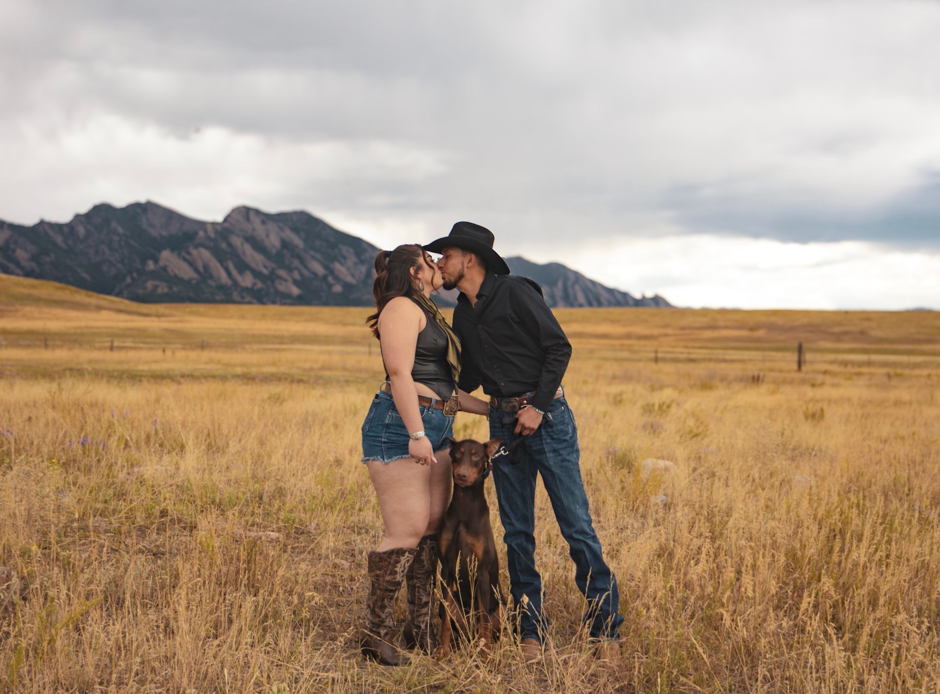 A man and a woman are kissing in a field with a dog.