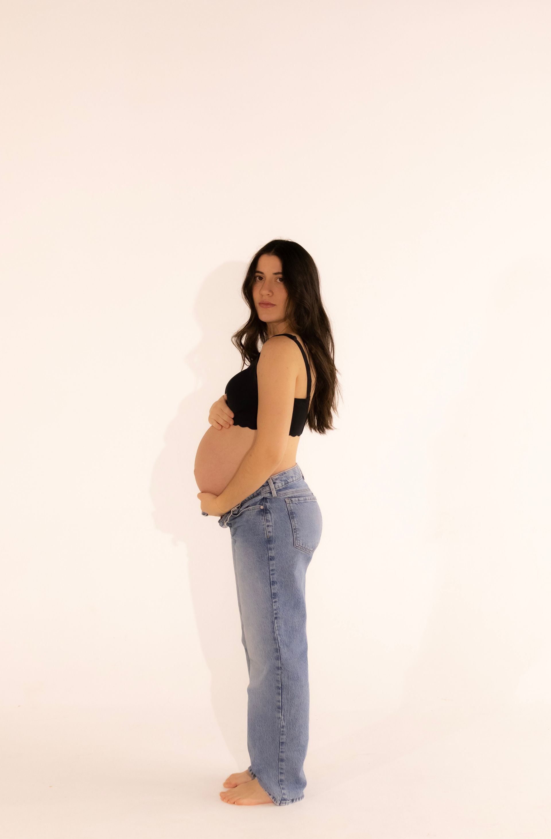 A pregnant woman is standing in front of a white wall holding her belly.