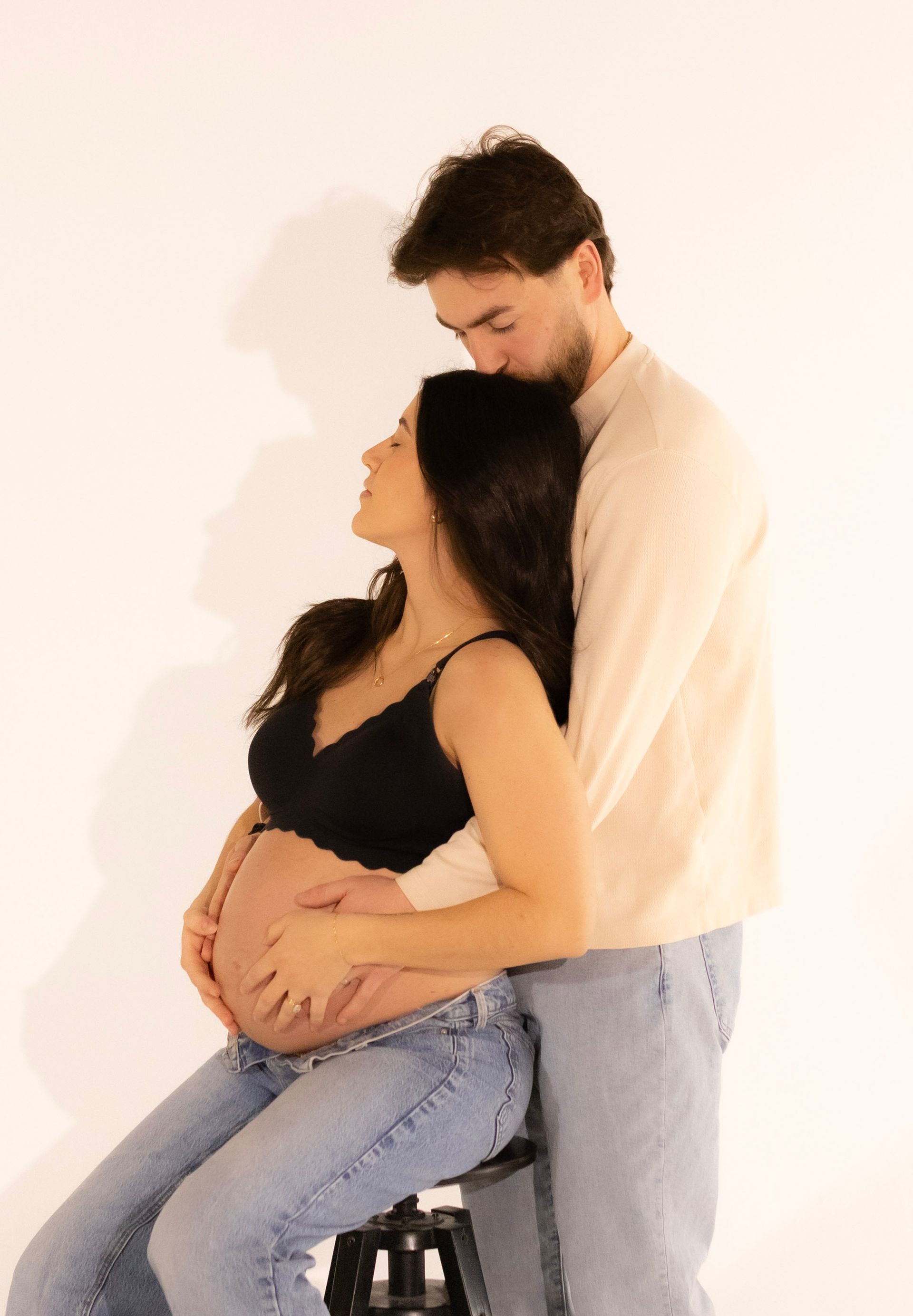 A man is hugging a pregnant woman who is sitting on a stool.