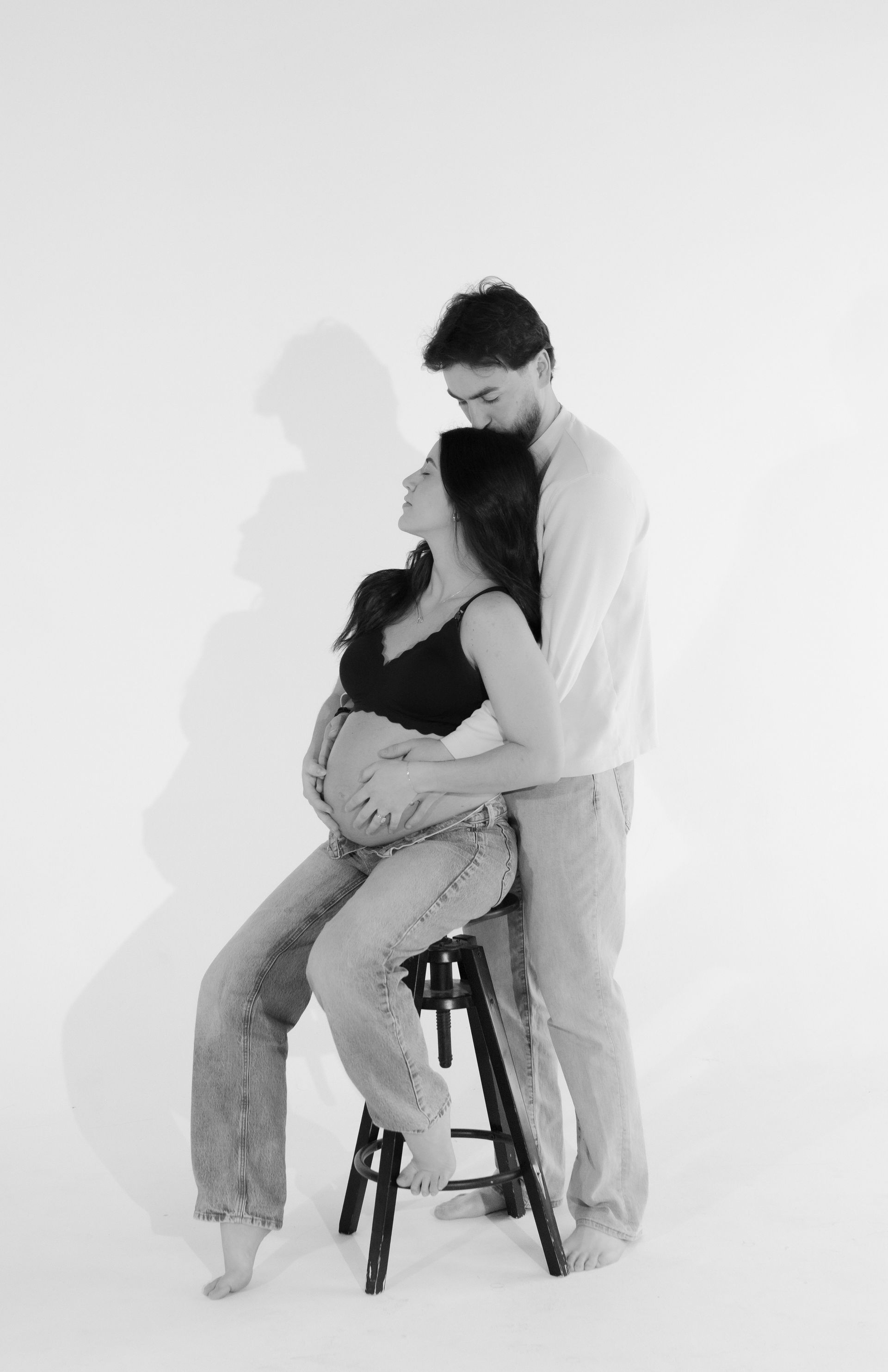 A man is hugging a pregnant woman sitting on a stool.