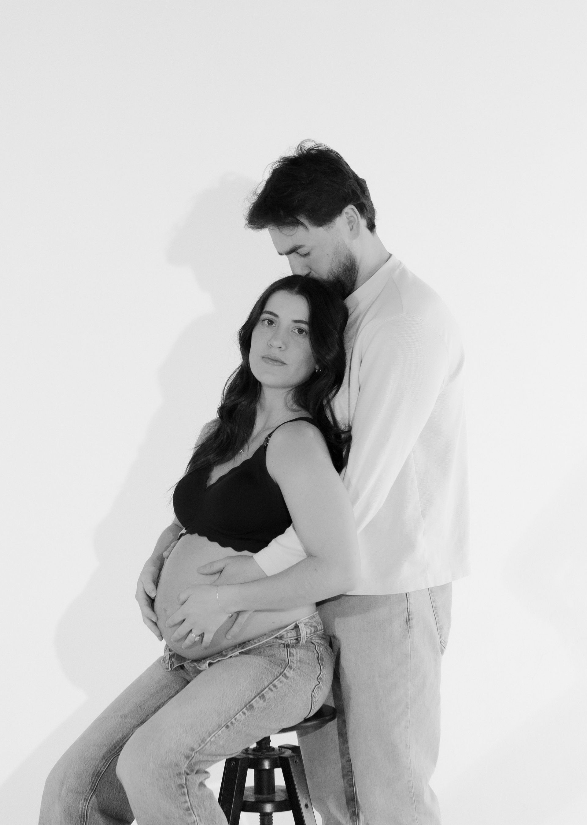A man is hugging a pregnant woman who is sitting on a stool.