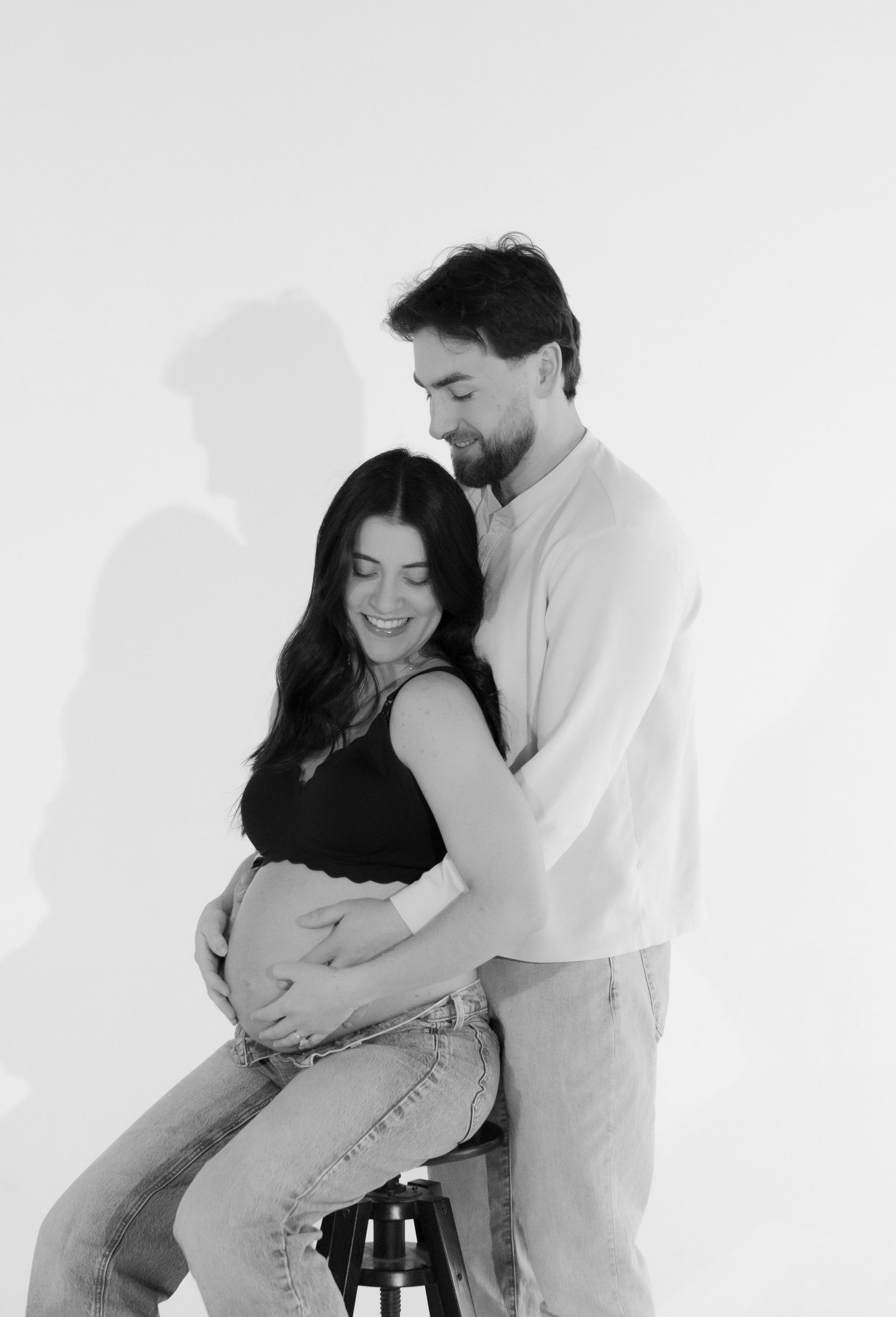 A man is hugging a pregnant woman who is sitting on a stool.