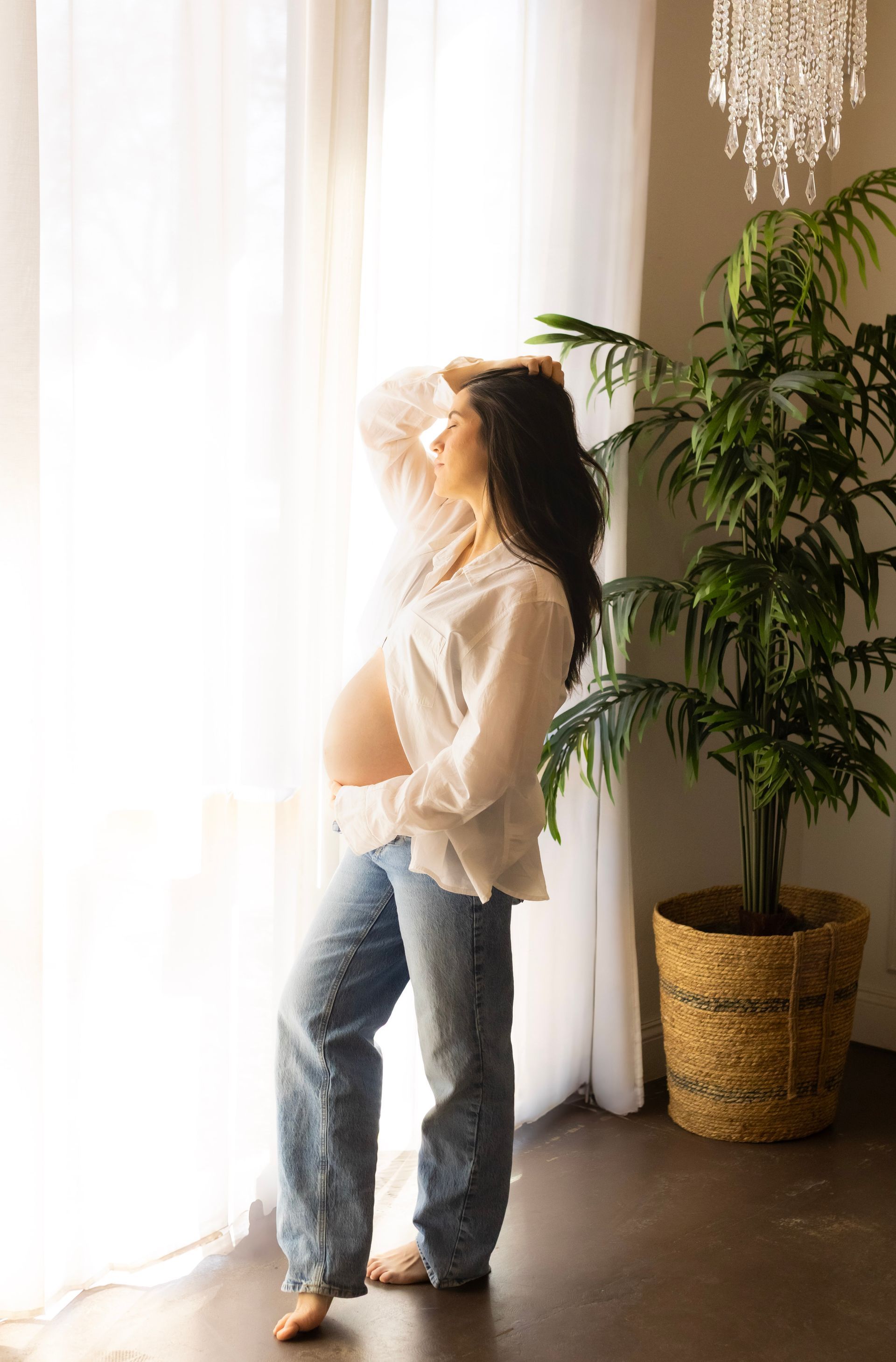 A pregnant woman in a white shirt and jeans is standing in front of a window.