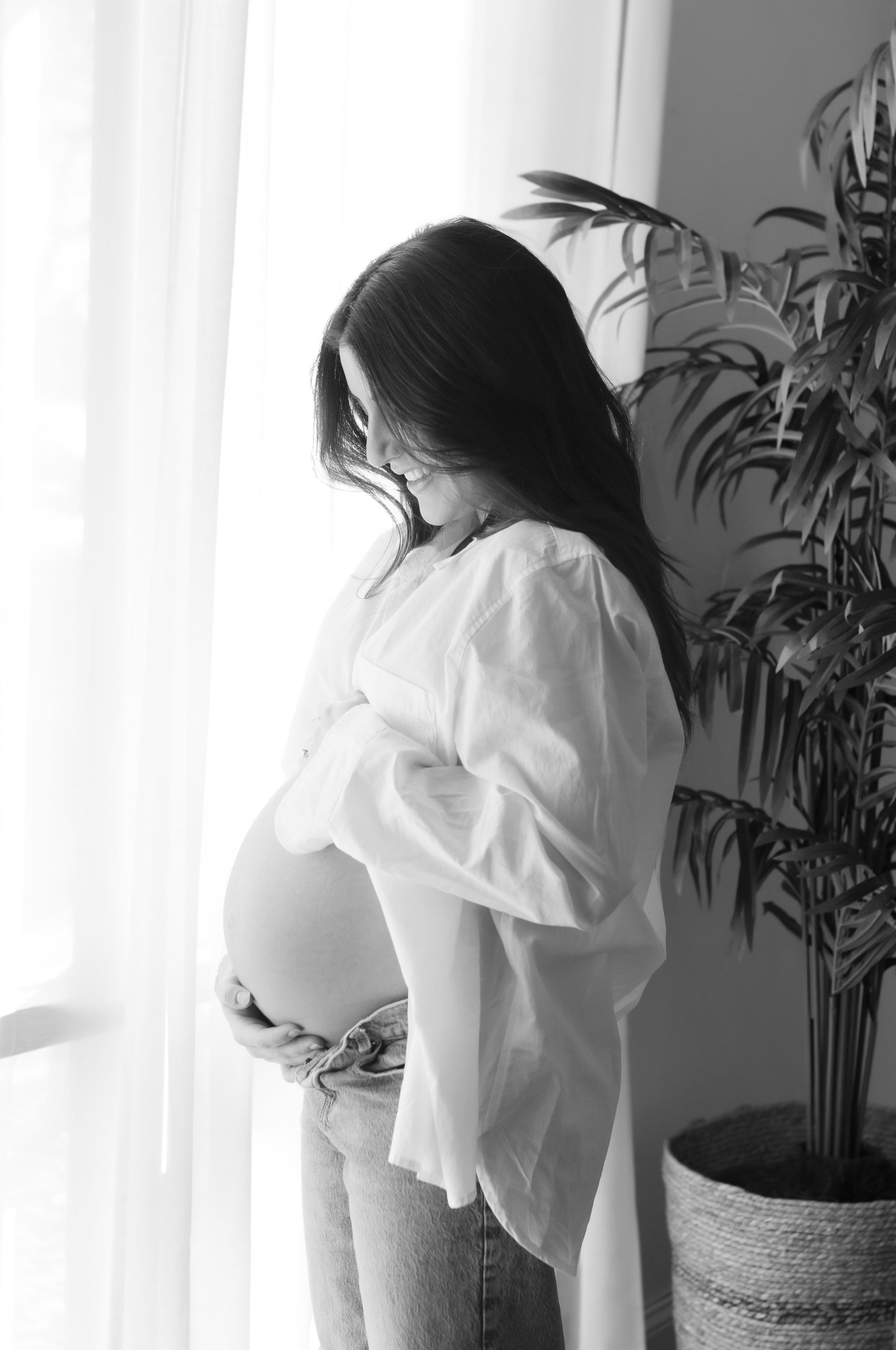A pregnant woman is standing in front of a window holding her belly.