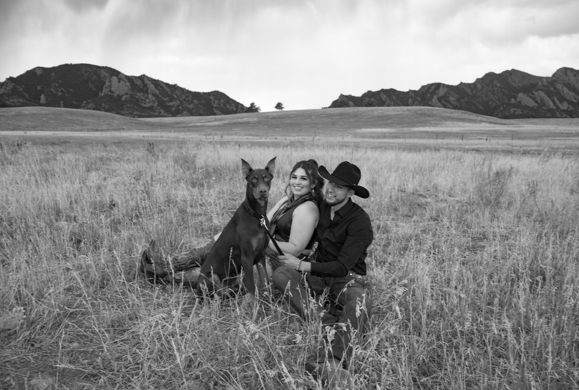 A black and white photo of a man and woman sitting in a field with a dog.