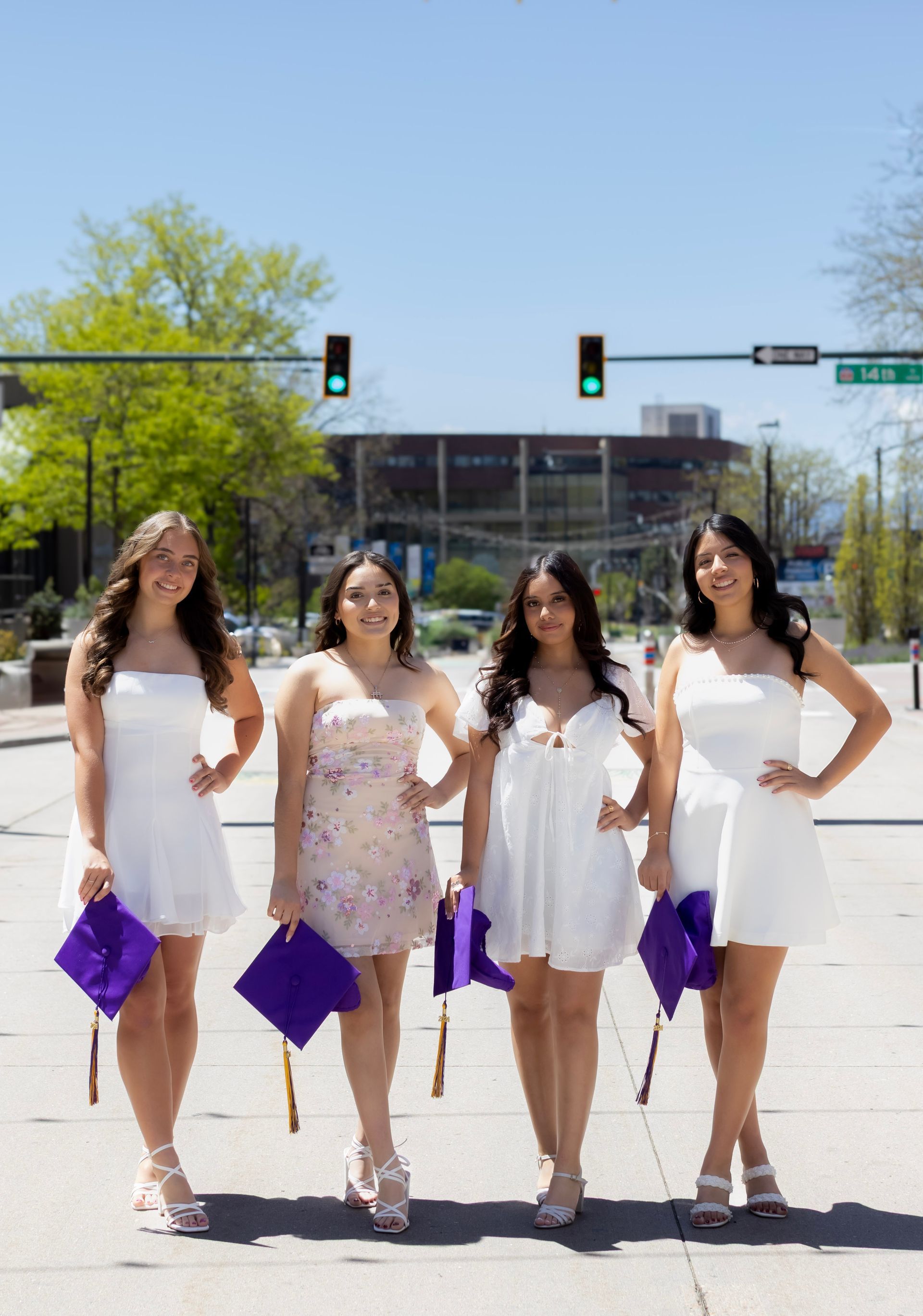 Four women in white dresses are standing on a sidewalk holding purple graduation caps.